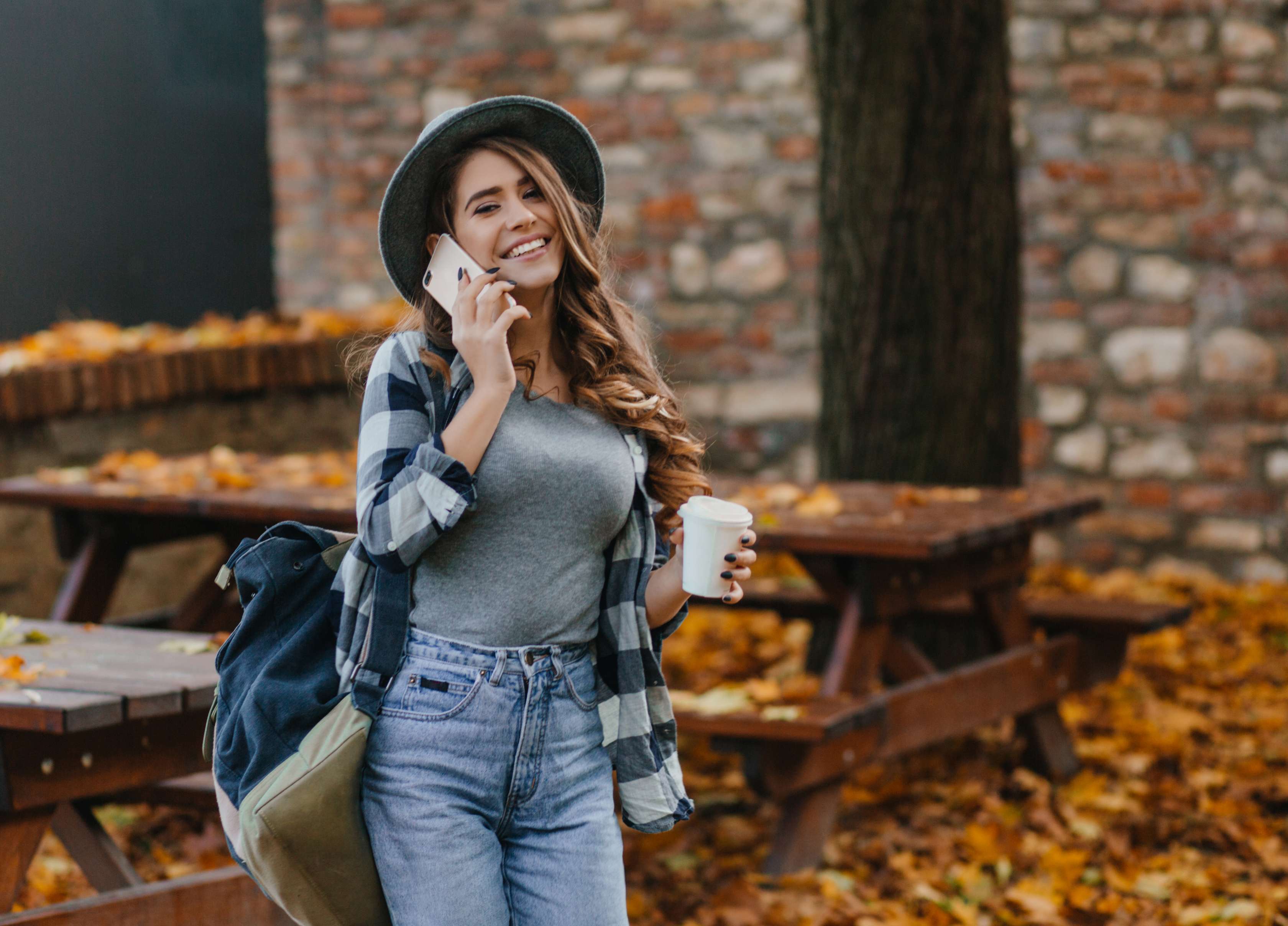 Worauf Sie bei einer günstigen Zahnzusatzversicherung achten sollten: Junge Frau mit Hut, Rucksack und Kaffeebecher in der Hand steht  vor einem Picknickplatz mit Herbstlaub und telefoniert.