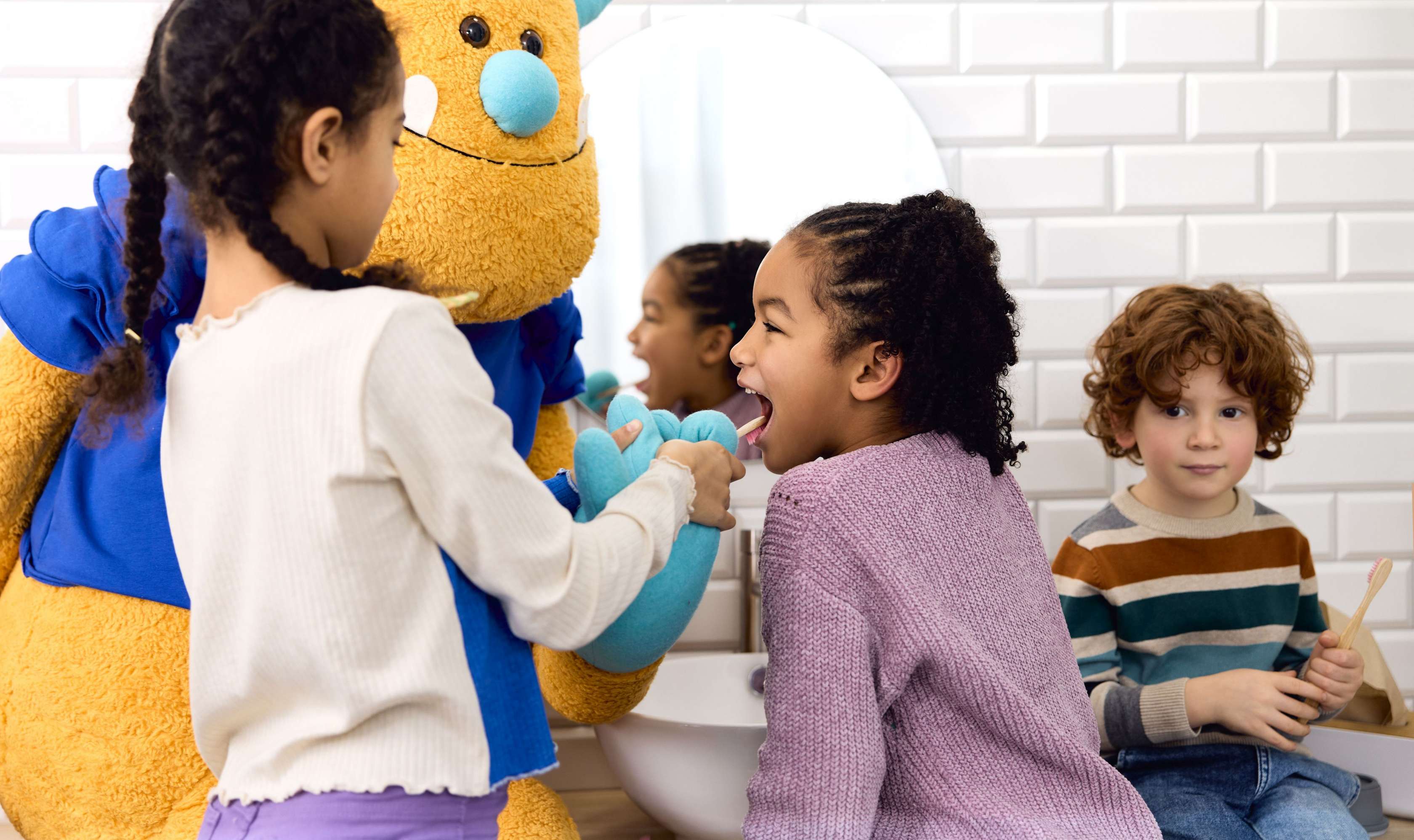 Zahnzusatzversicherung für Kinder: Kleine Kinder stehen mit einem großen gelben Plüschmonster zusammen im Bad und putzen sich die Zähne