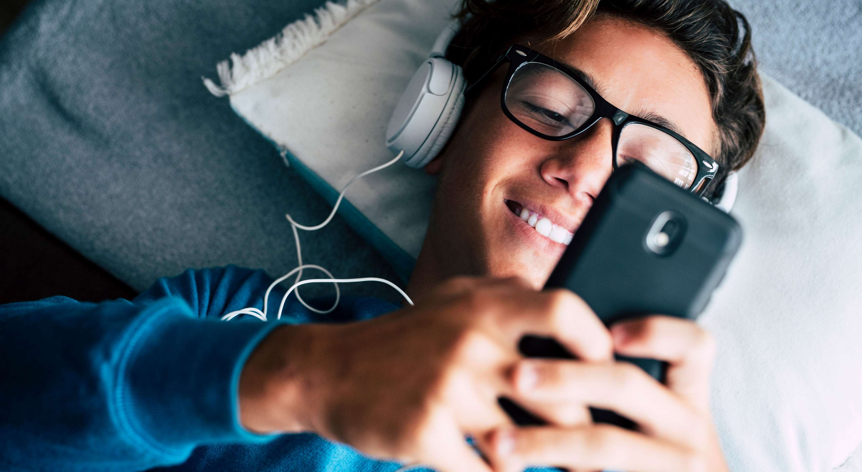 Ein junger Mann liegt mit Kopfhörern auf dem Fußboden und sieht lächelns in sein Handy