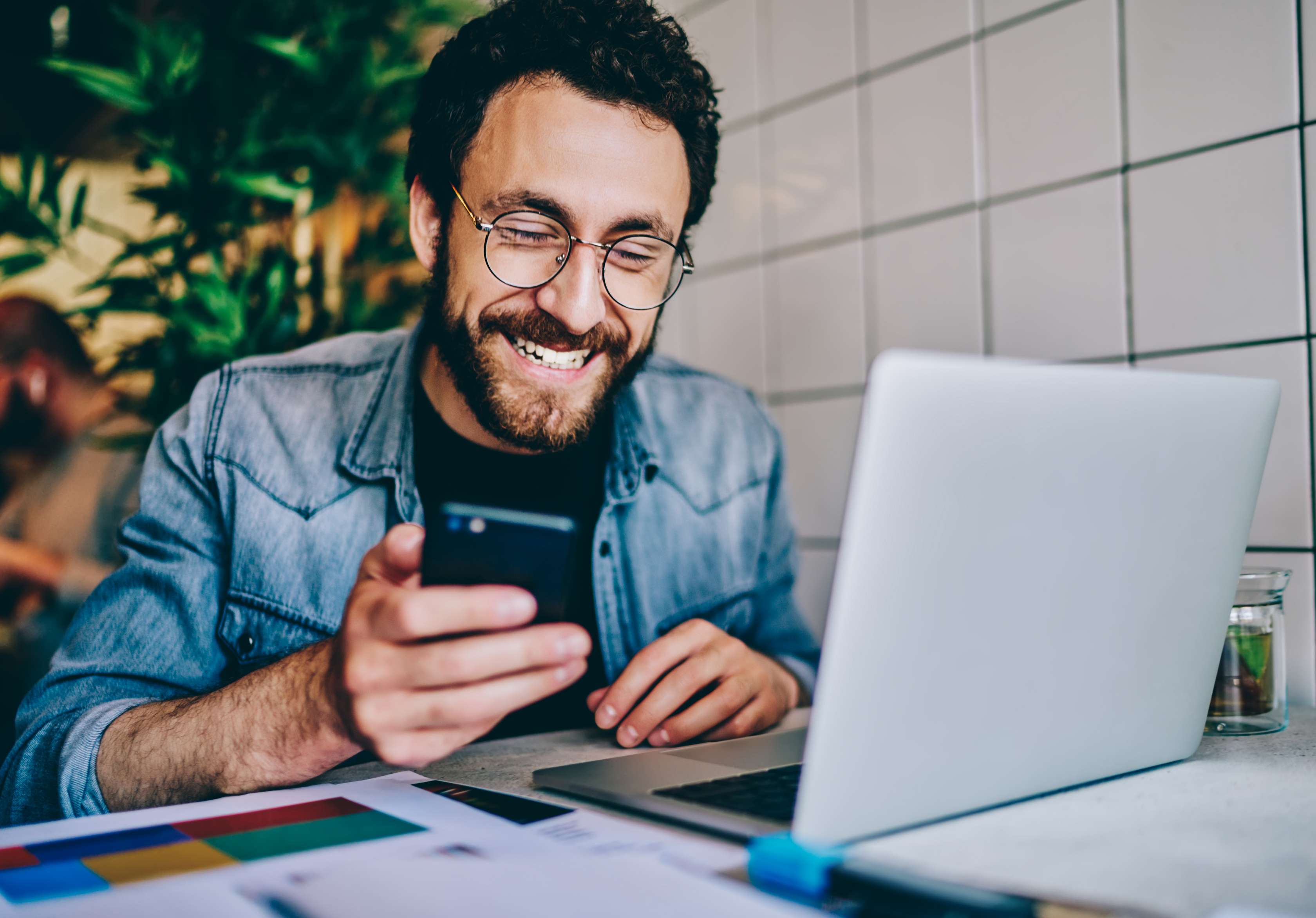 Zahnzusatzversicherung – wie Sie die ZZV richtig wechseln: Mann mit Brille und Smartphone in der Hand sitzt lächelnd vor einem Laptop und Unterlagen.