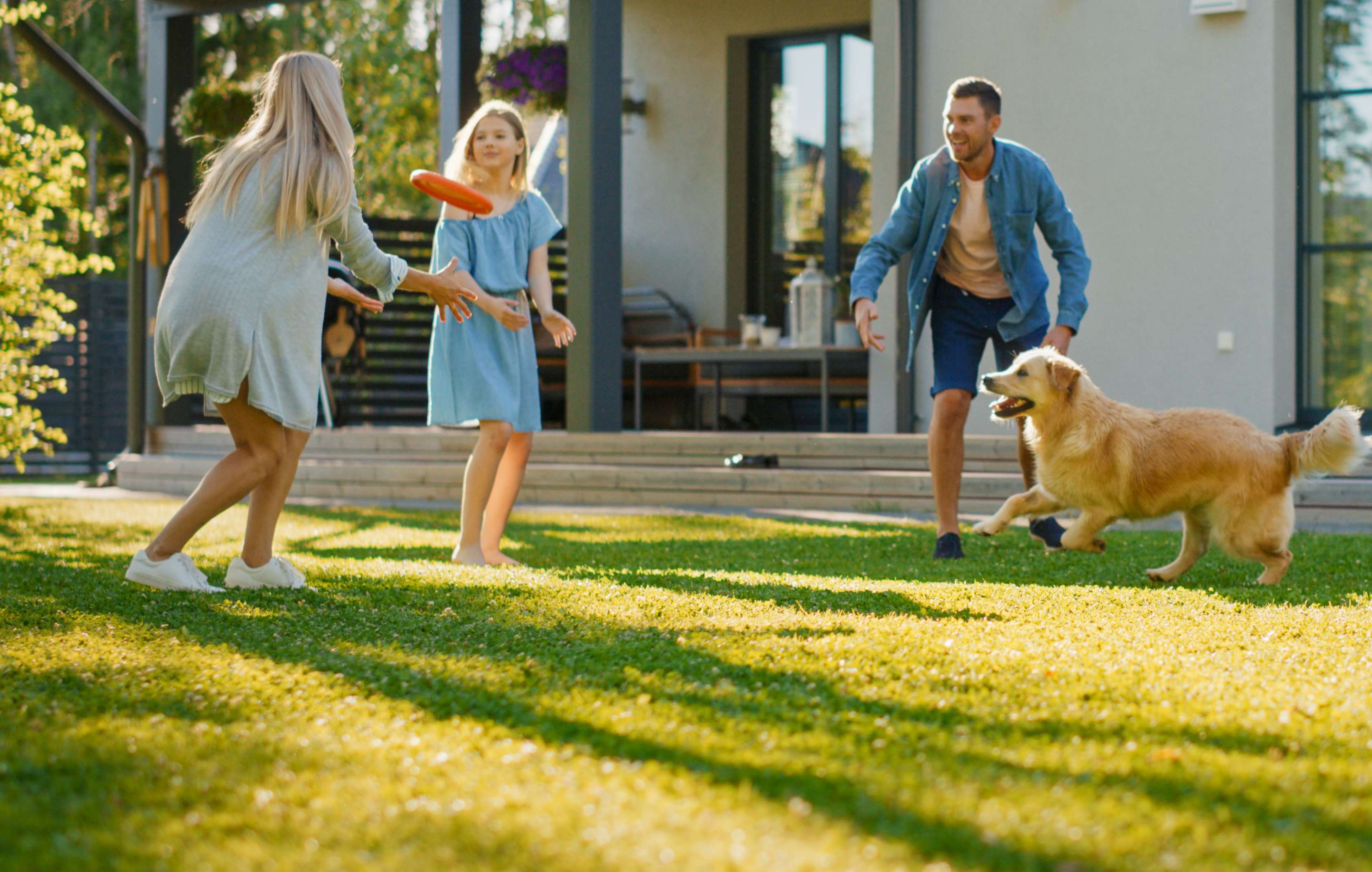 Allianz Baufinanzierung: Eltern spielen mit zwei Kindern und Hund Frisbee vor der Terrasse ihres Hauses