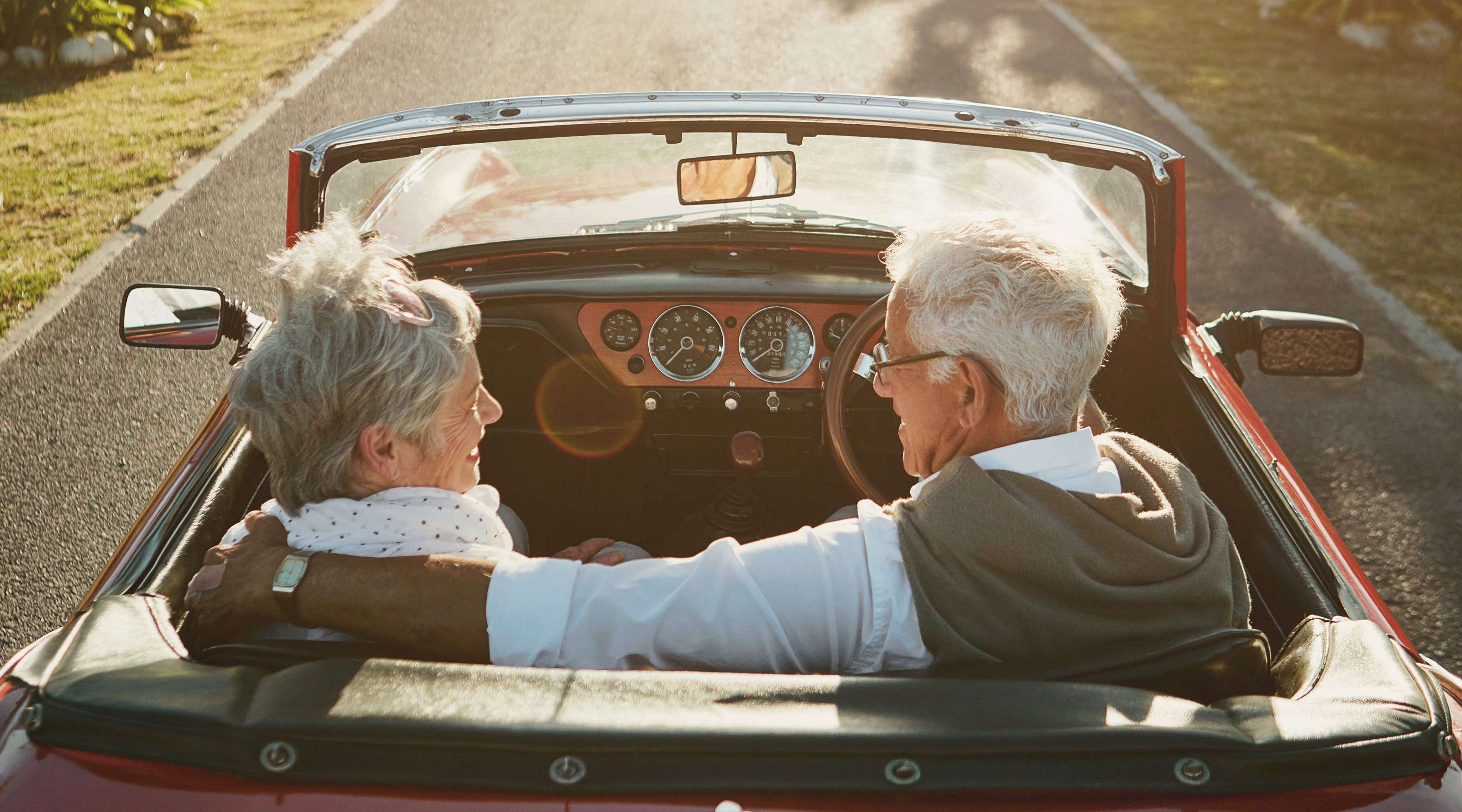 Allianz BestAger Finanzierung - Ein älteres Paar sitzt in einem roten Auto auf dem Weg in die Ferne und lächelt sich an.