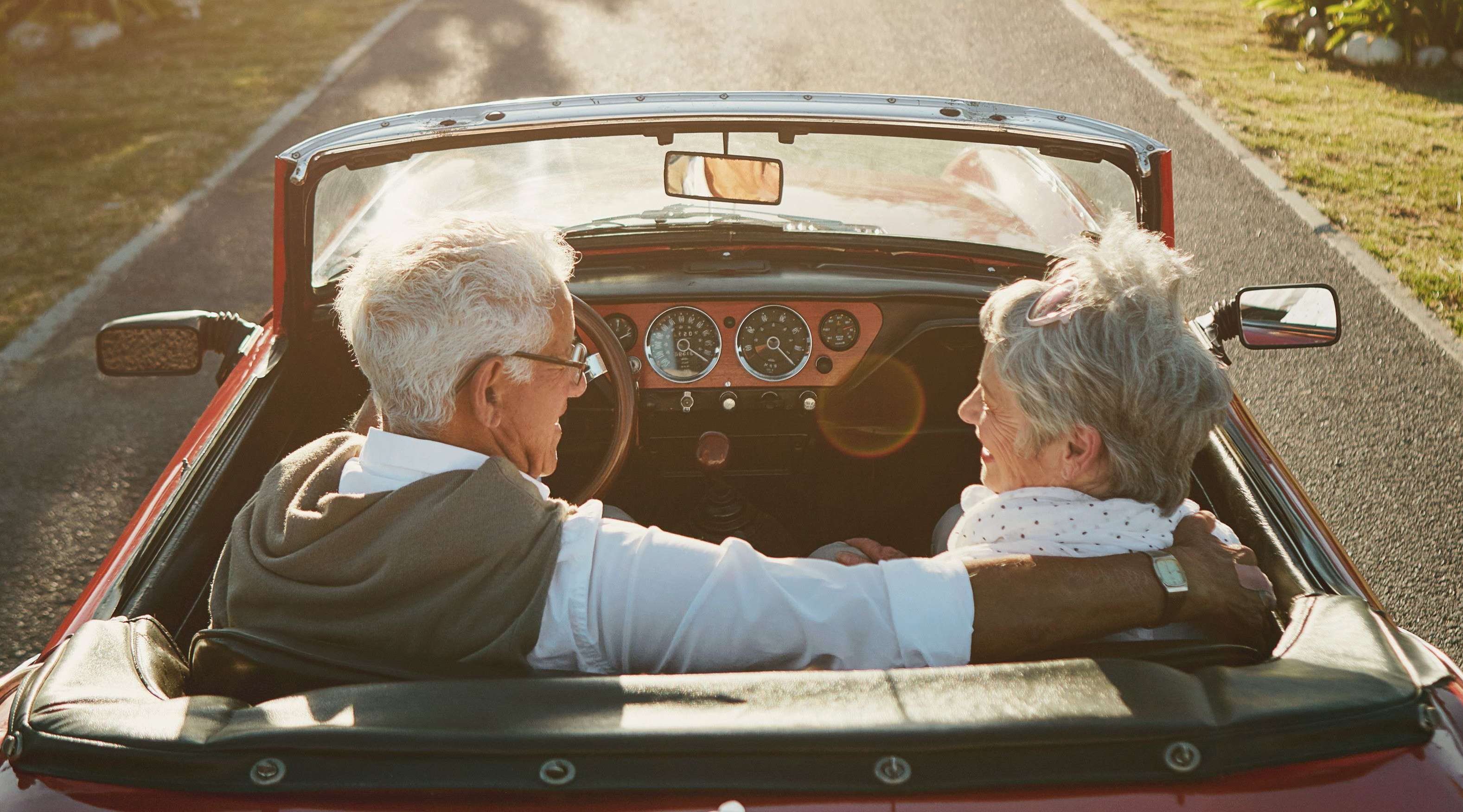 Allianz BestAger Finanzierung - Ein älteres Paar sitzt in einem roten Auto auf dem Weg in die Ferne und lächelt sich an.