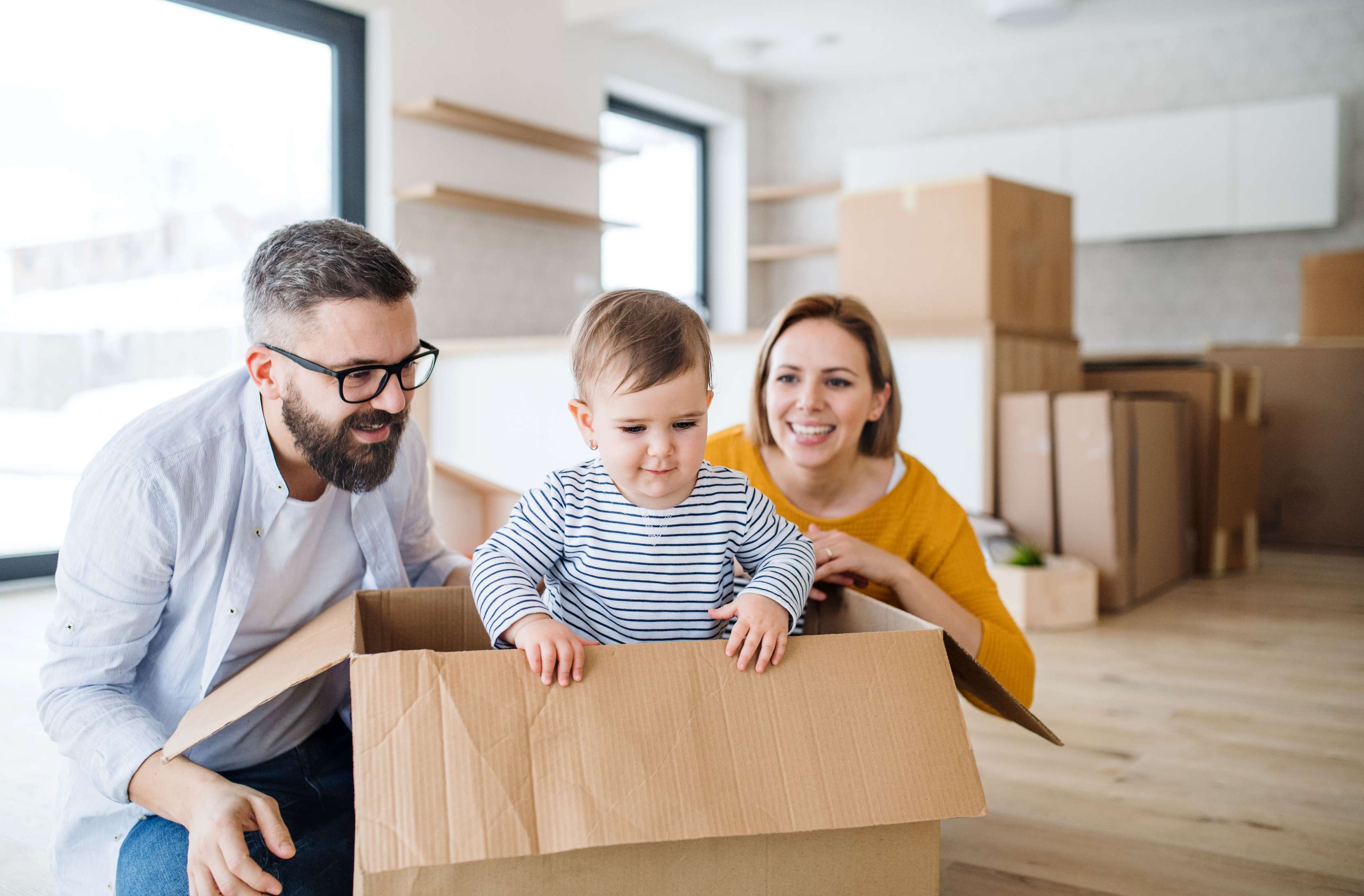 Allianz Immobilienförderung: Ein Paar sitzt gemeinsam in ihrem neuen Haus auf dem Boden. Zwischen ihnen steht ein offener Umzugskarton, in dem ihr Kleinkind steht.