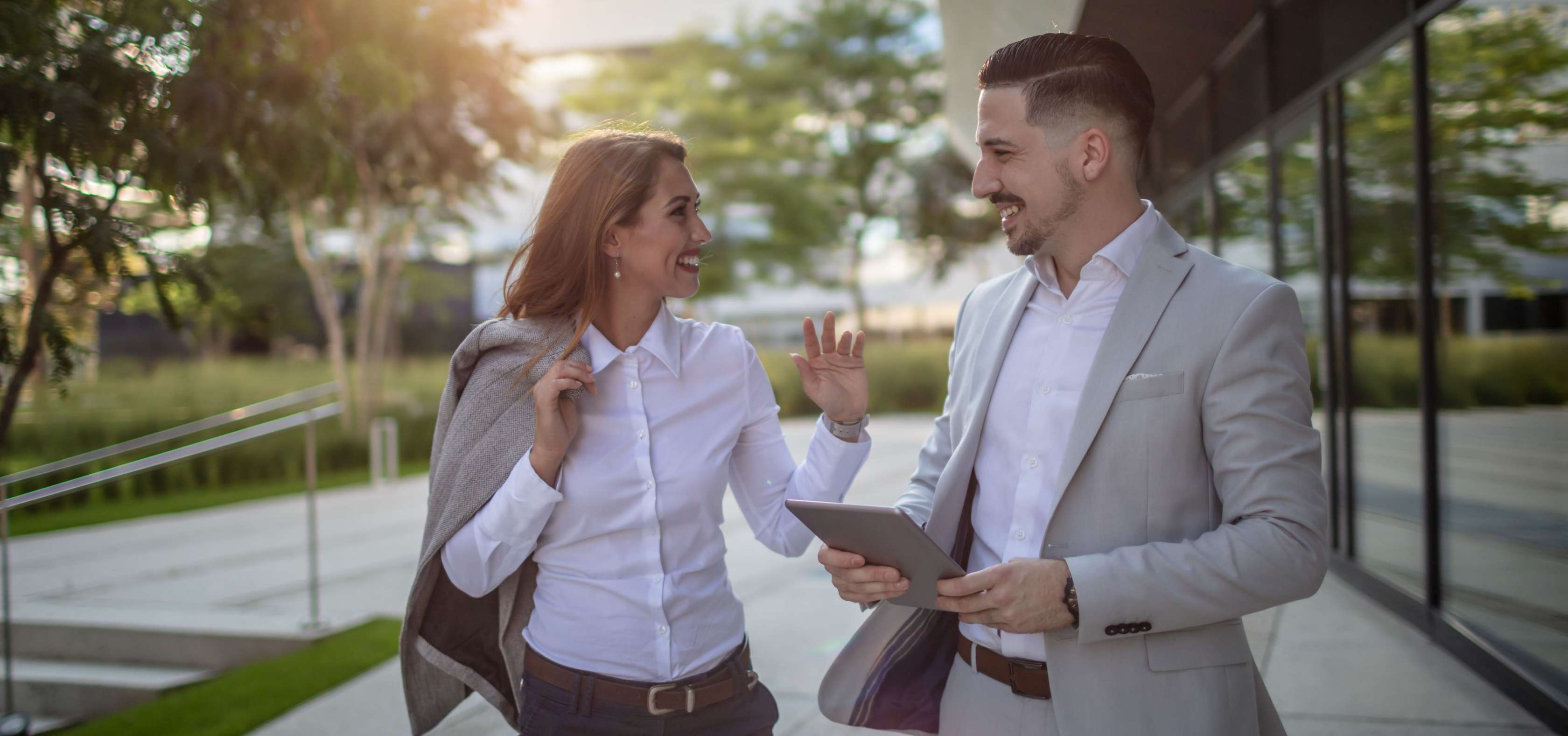 Allianz Teilungserklärung - Ein Mann und eine Frau in Business-Kleidung gehen draußen und unterhalten sich lächelnd. Der Mann hält ein Tablet in der Hand.