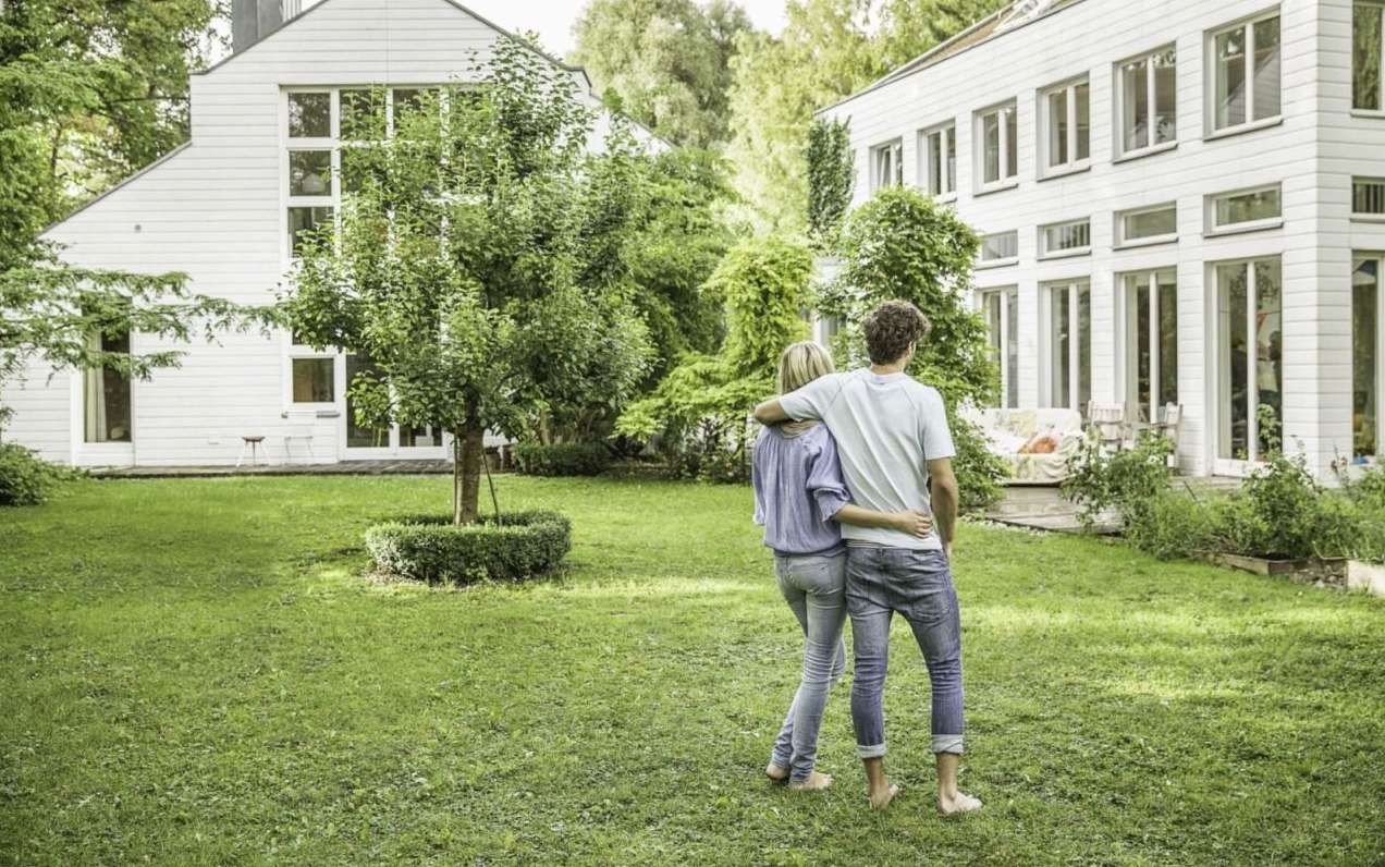 Allianz Immobilien Wertermittlung - Ein Paar steht Arm in Arm in einem grünen Garten und blickt auf zwei weiße Häuser.
