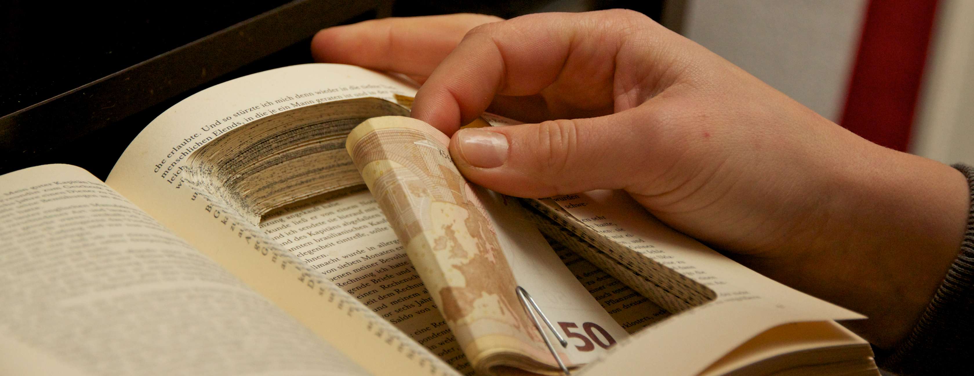 Nahaufnahme einer Frauenhand, die ein Bündel 50-Euro-Scheine in ein aufgeschlagenes Buch legt, das als Geheimversteck präpariert wurde: In der Mitte der rechten Seiten des Buches sieht man eine in das Papier geschnittene Aussparung zum Verstecken des Geldes.