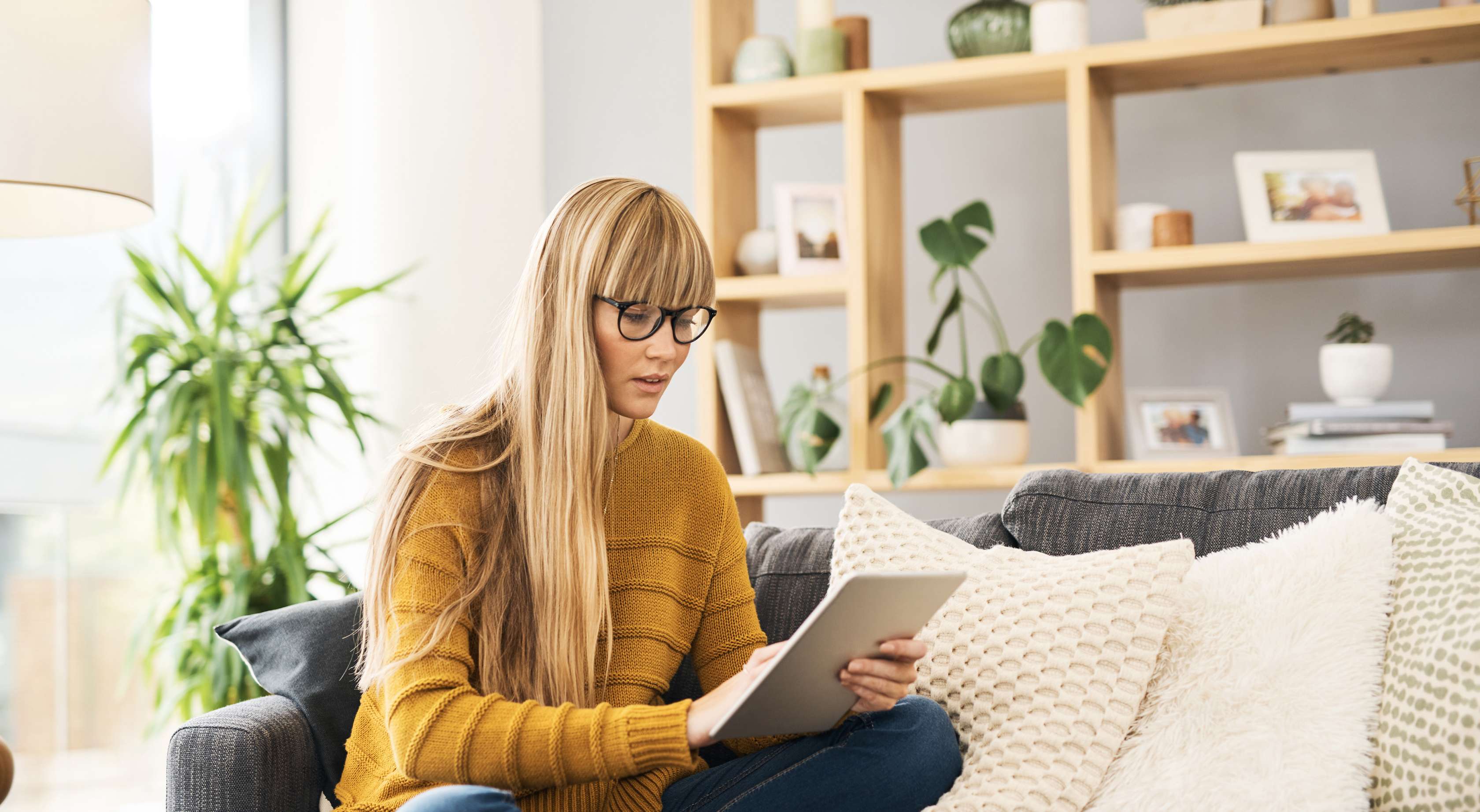 Junge blonde Frau im gelben Pullover sitzt auf Couch und schaut in ihr Tablet