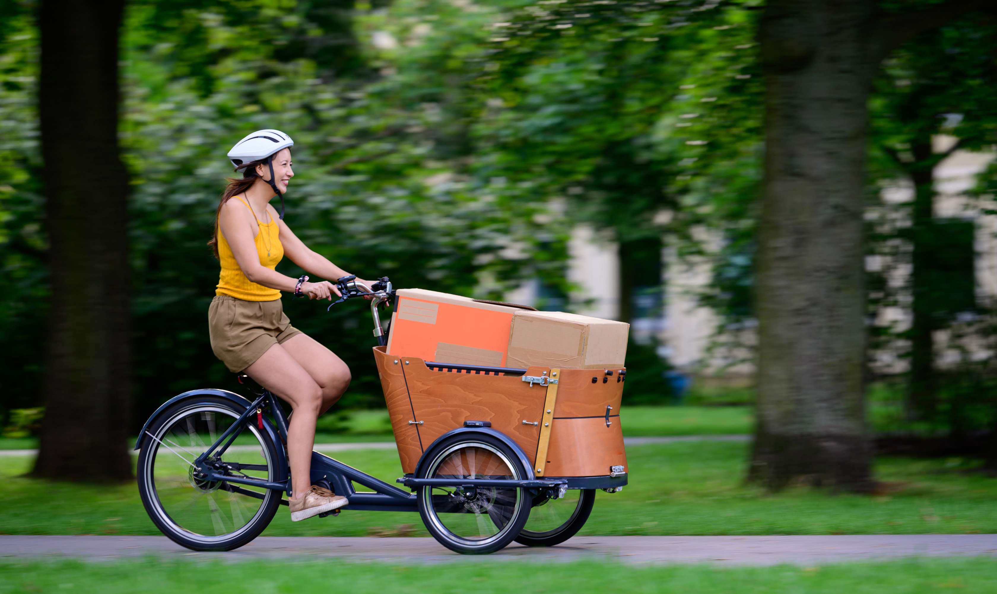 Lastenrd versichern: Eine Frau fährt mit einem Lastenrad und einem Paket durch einen Park