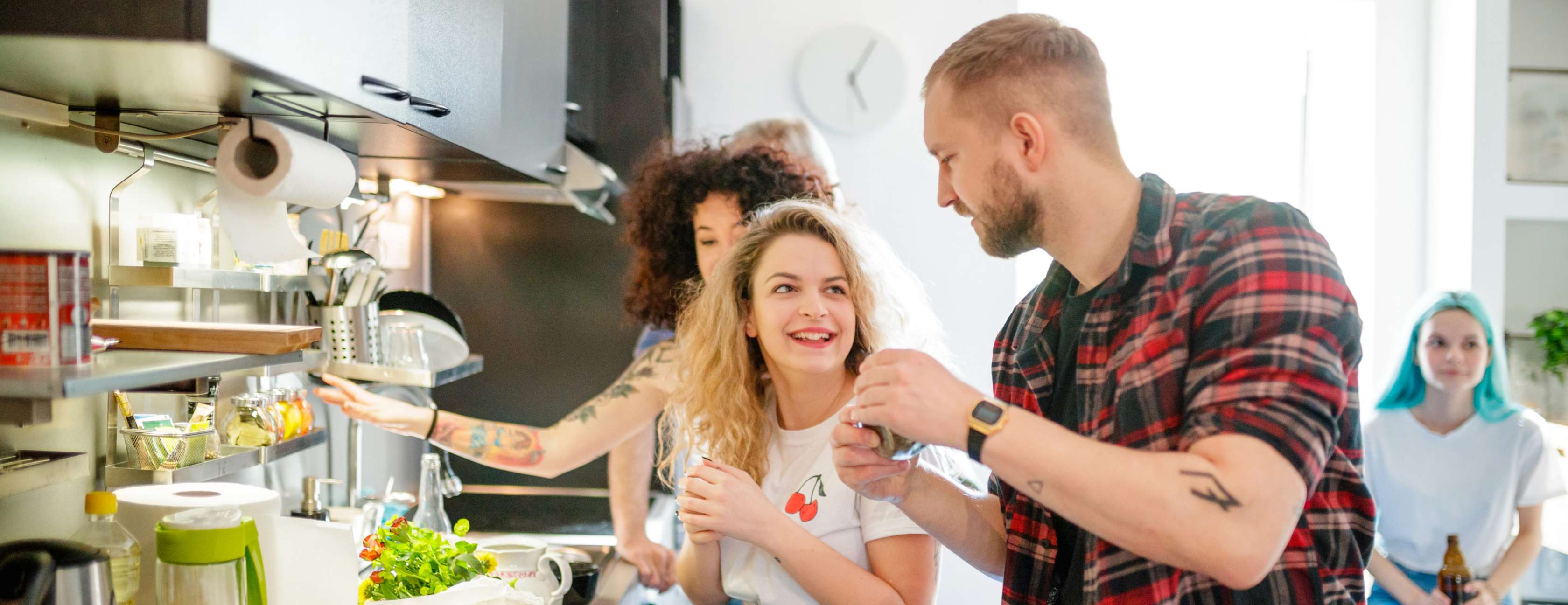Zwei junge Frauen und ein junger Mann stehend zusammen lachend in einer WG-Küche bei der gemeinsamen Essensvorbereitung.