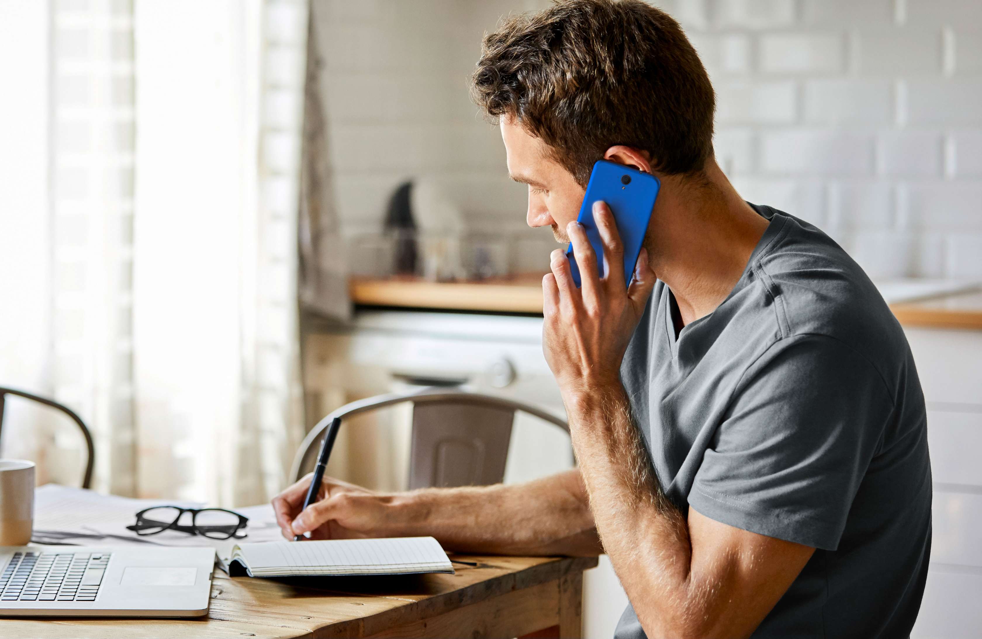 Ein junger Mann sitzt in einer hell eingerichteten Wohnung an einem Tisch mit Laptop, telefoniert und notiert sich etwas in ein Notizbuch – Symbolbild Rechtsschutzversicherung