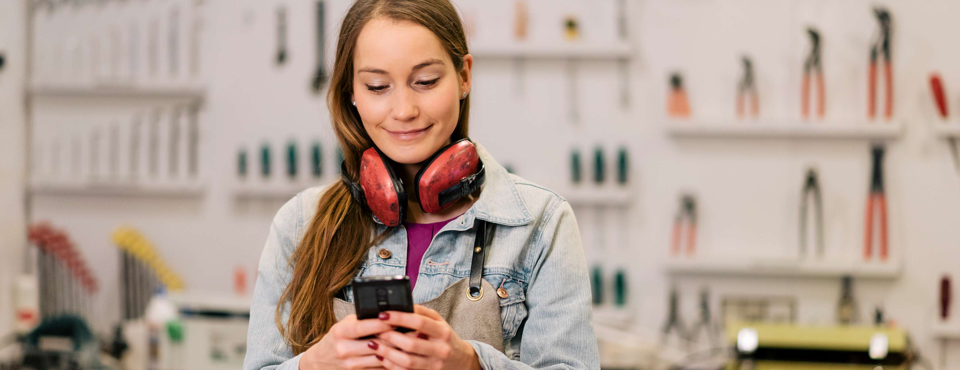 Allianz - Smartphone Nutzung am Arbeitsplatz: Junge Frau im Monteursanzug bedient Handy in einer Werkstatt