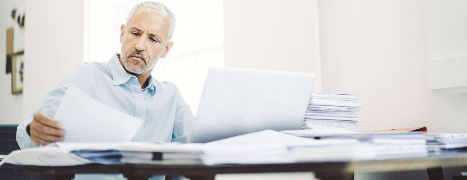 Ein Mann schaut in seine Unterlagen, während er an einem Schreibtisch sitzt, auf dem Dokumente verstreut sind und ein Laptop steht