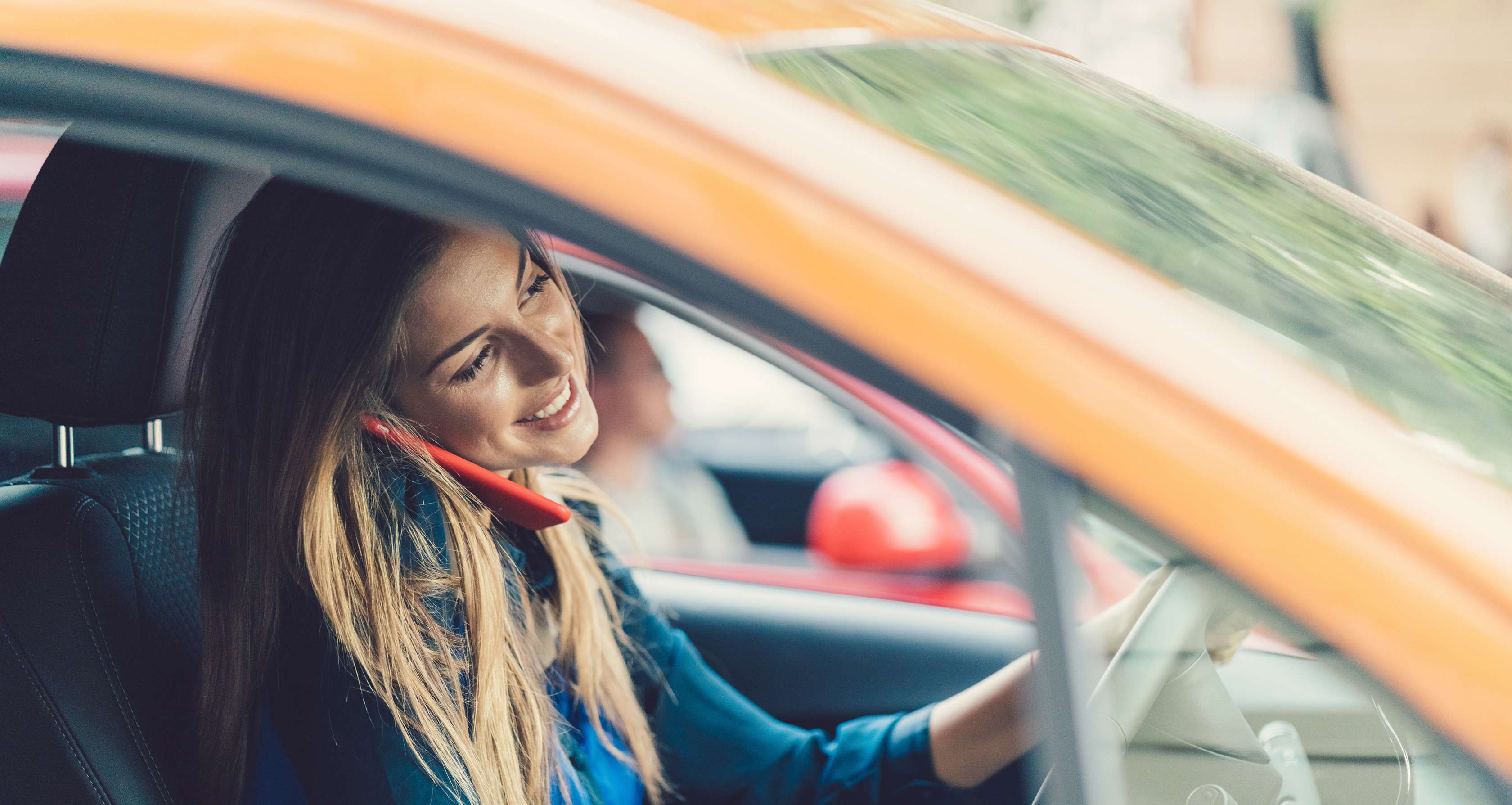 Eine blonde, langhaarige Frau hat ein rotes Smartphone an der Schulter eingeklemmt und telefoniert im Auto