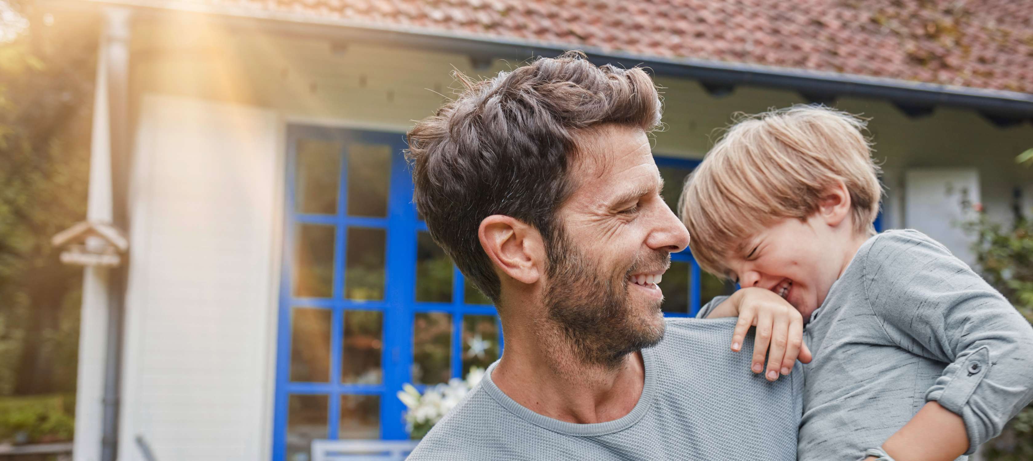 Wohngebäudeversicherung: Vater steht mit Sohn auf dem Arm glücklich vor seinem Haus
