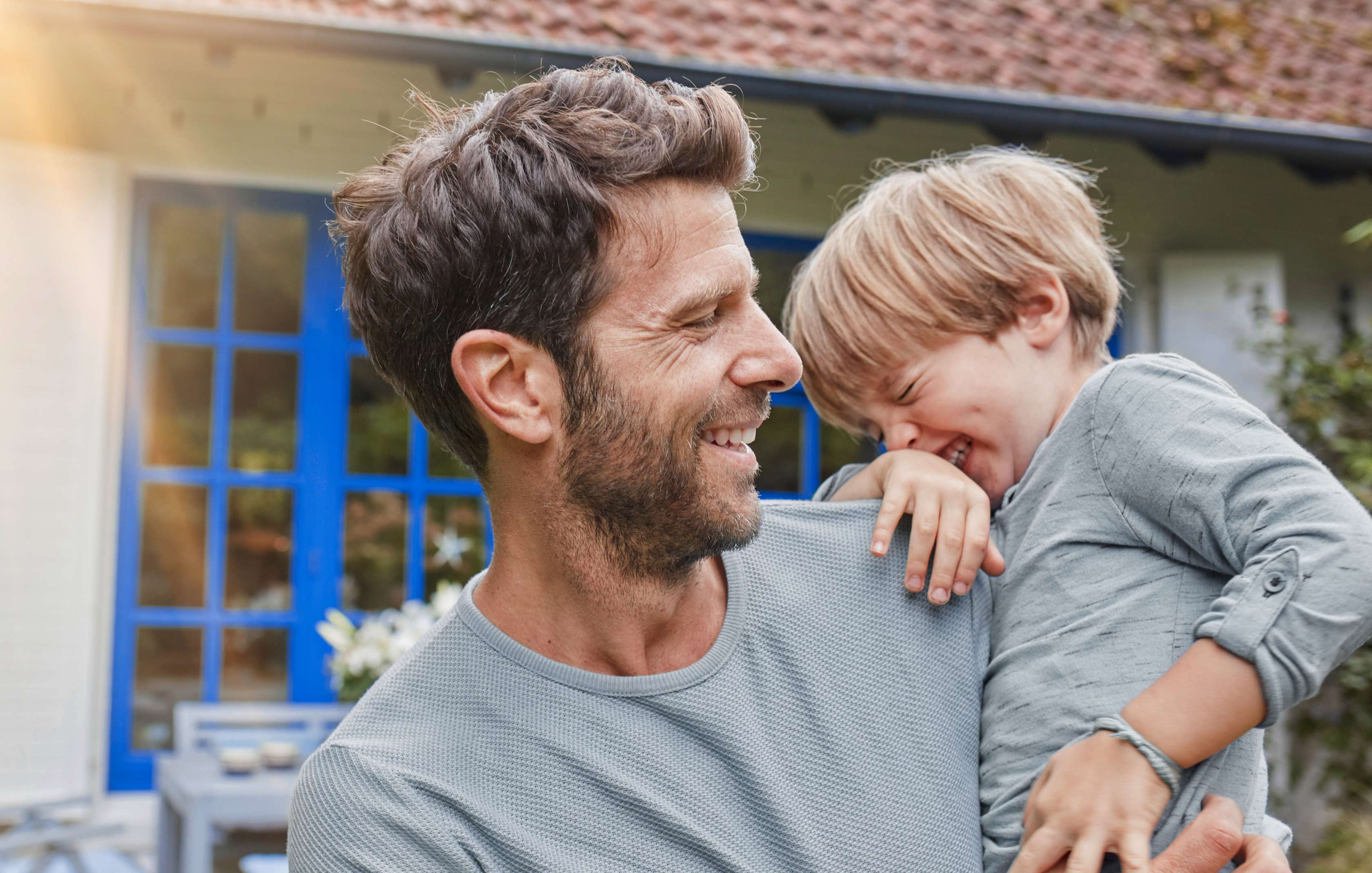 Glücklich lachender Vater steht mit seinem kleinem Sohn auf dem Arm im Garten vor seinem Haus – Symbolbild Wohngebäudeversicherung 