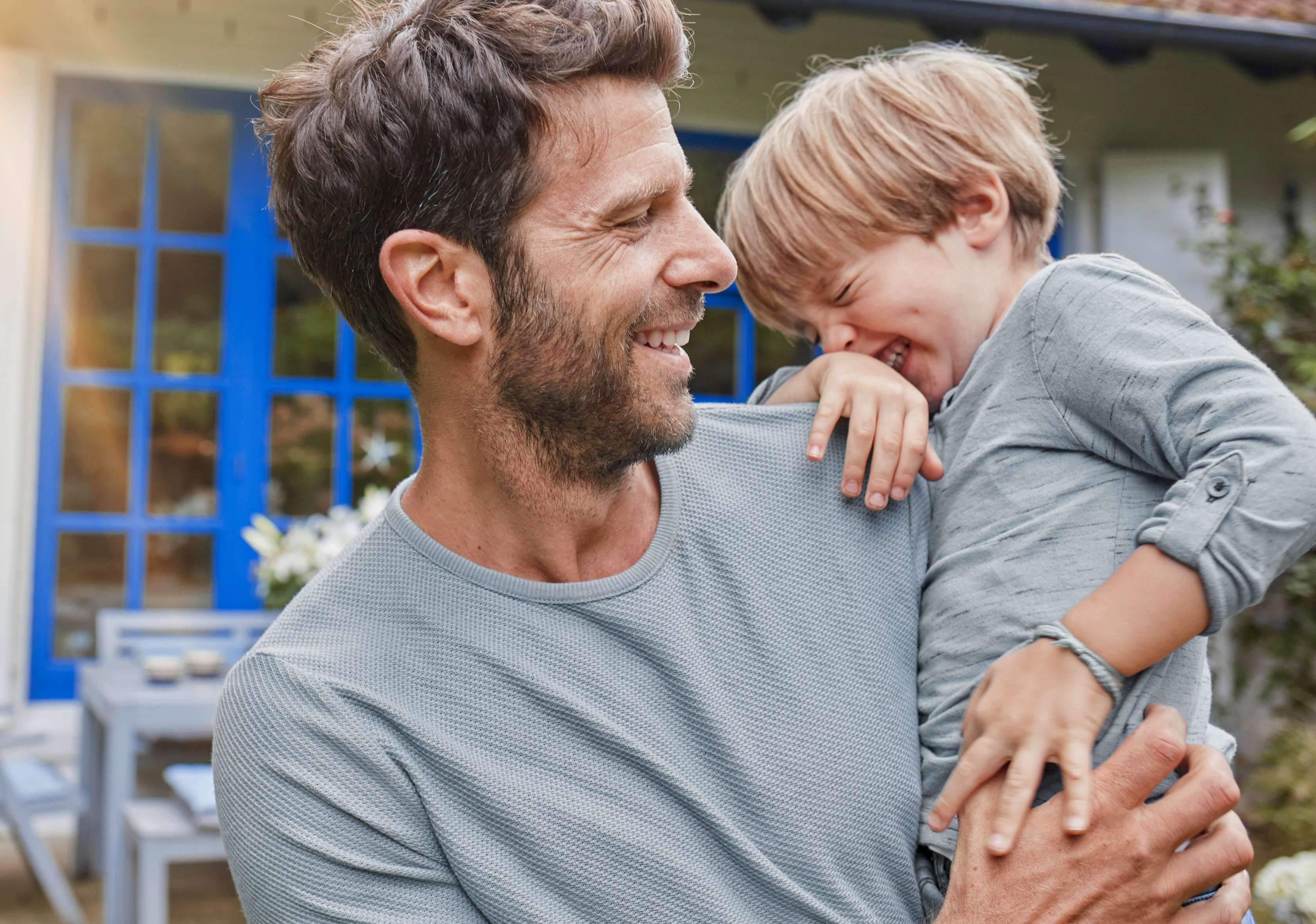 Glücklich lachender Vater steht mit seinem kleinem Sohn auf dem Arm im Garten vor seinem Haus – Symbolbild Wohngebäudeversicherung 