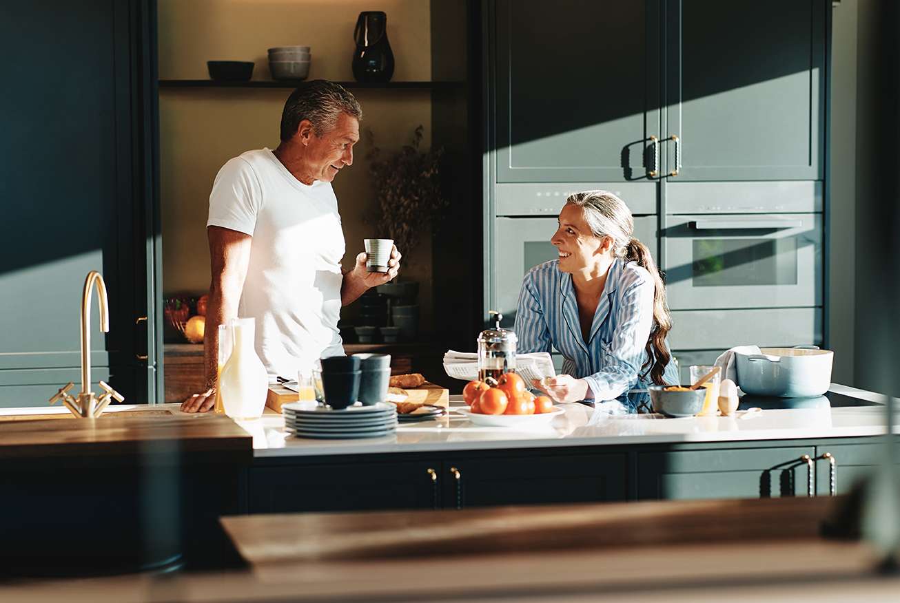 Zwei Erwachsene in einer hellen Küche: Eine Person steht mit einer Tasse am Küchentisch, die andere sitzt davor. Auf dem Tisch liegen Tomaten, Brot und eine Kaffeekanne.