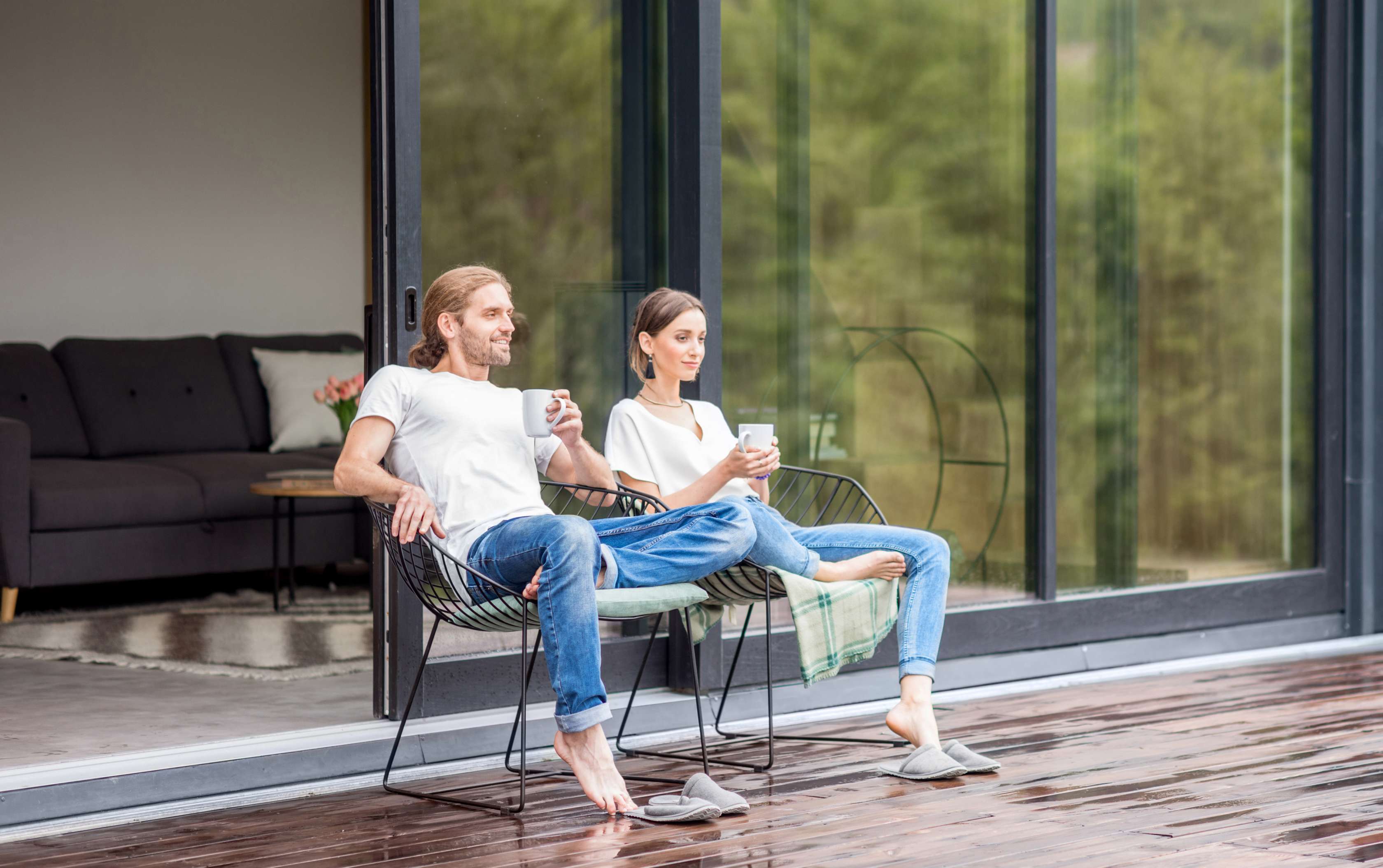 Haus-Haftpflichtversicherung: Ein Paar sitzt entspannt mit einer Tasse Kaffee in der Hand auf der Terrasse vor seinem Haus 