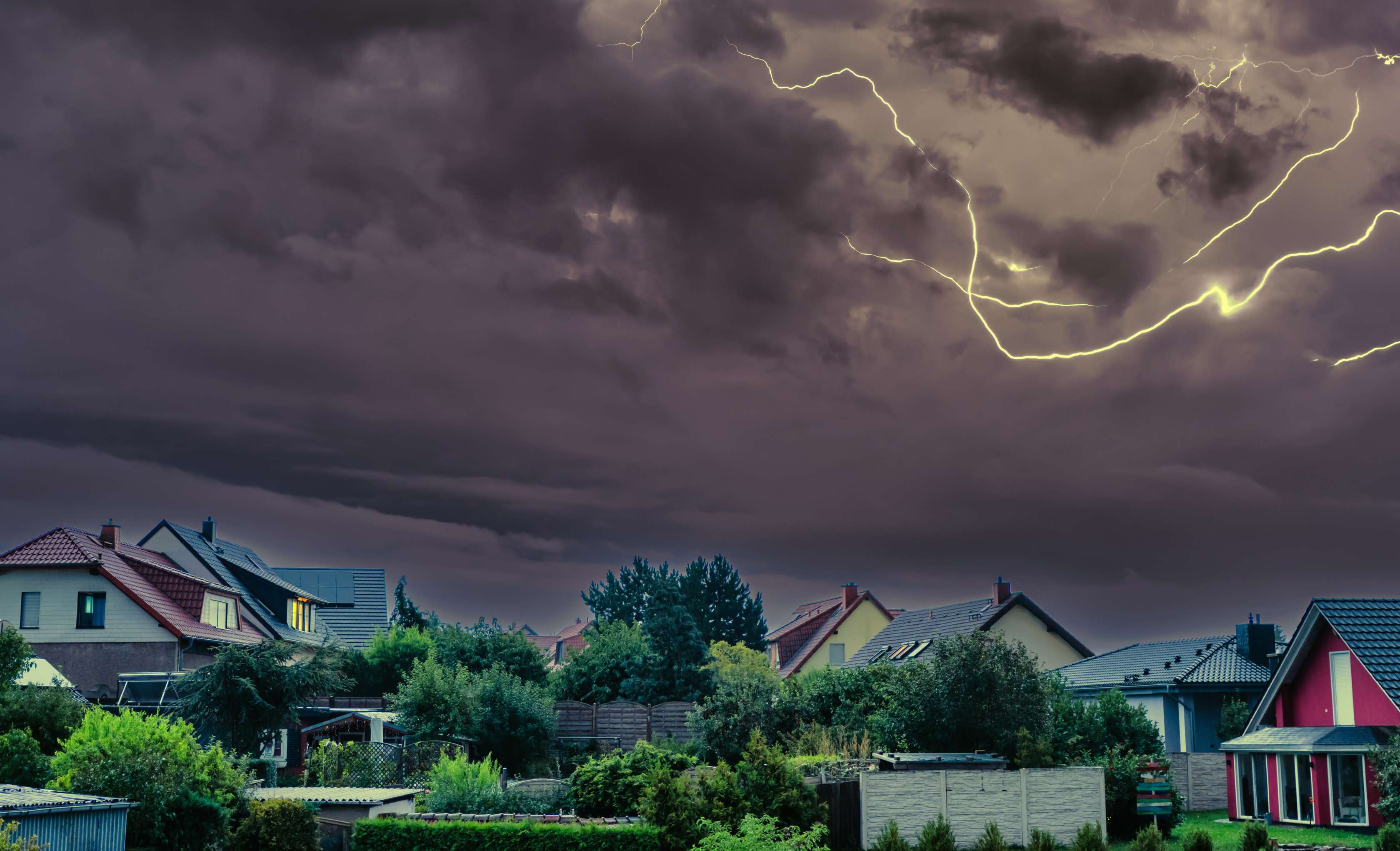 Sturmschaden Versicherung: Blitze und dunkle Gewitterwolken über einer Siedlung mit mehreren Einfamilienhäusern