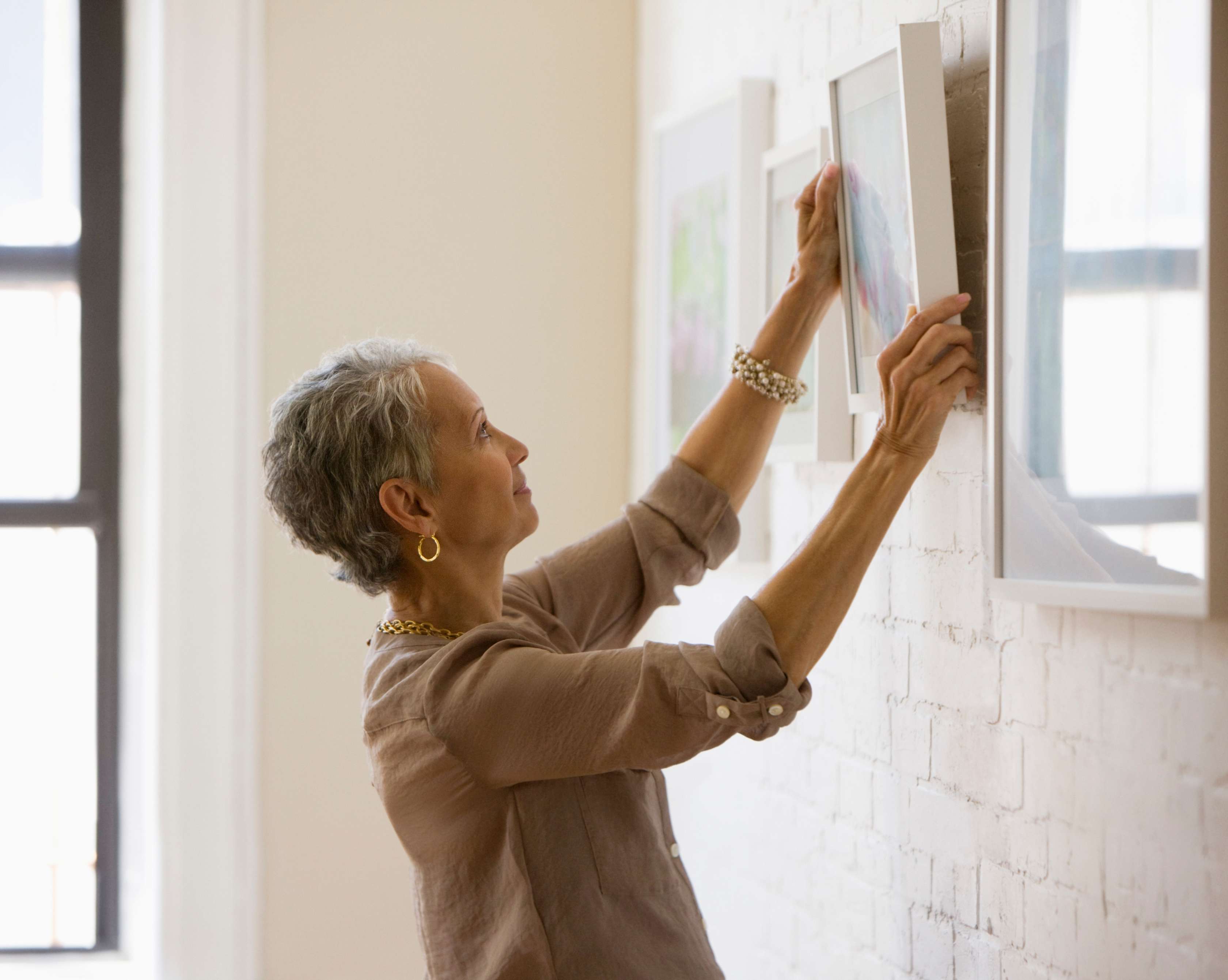 Allianz - Kunstversicherung: Erfahrene Frau hängt gerahmtes Bild an Zimmerwand