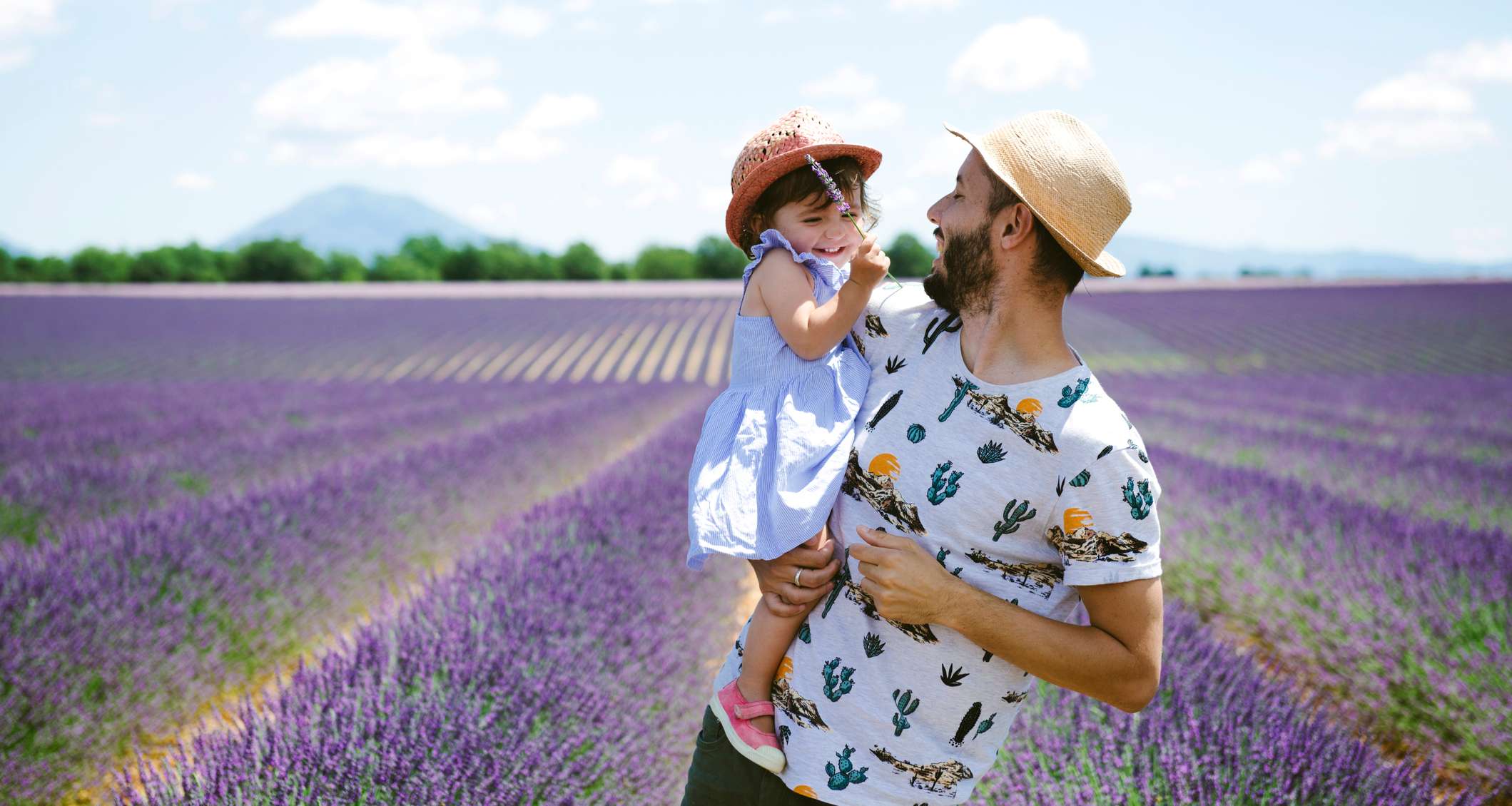 Lachender Vater mit kleiner Tocjter auf dem Arm steht in einem Lavendelfeld. Beide haben Strohhüte auf dem Kopf.