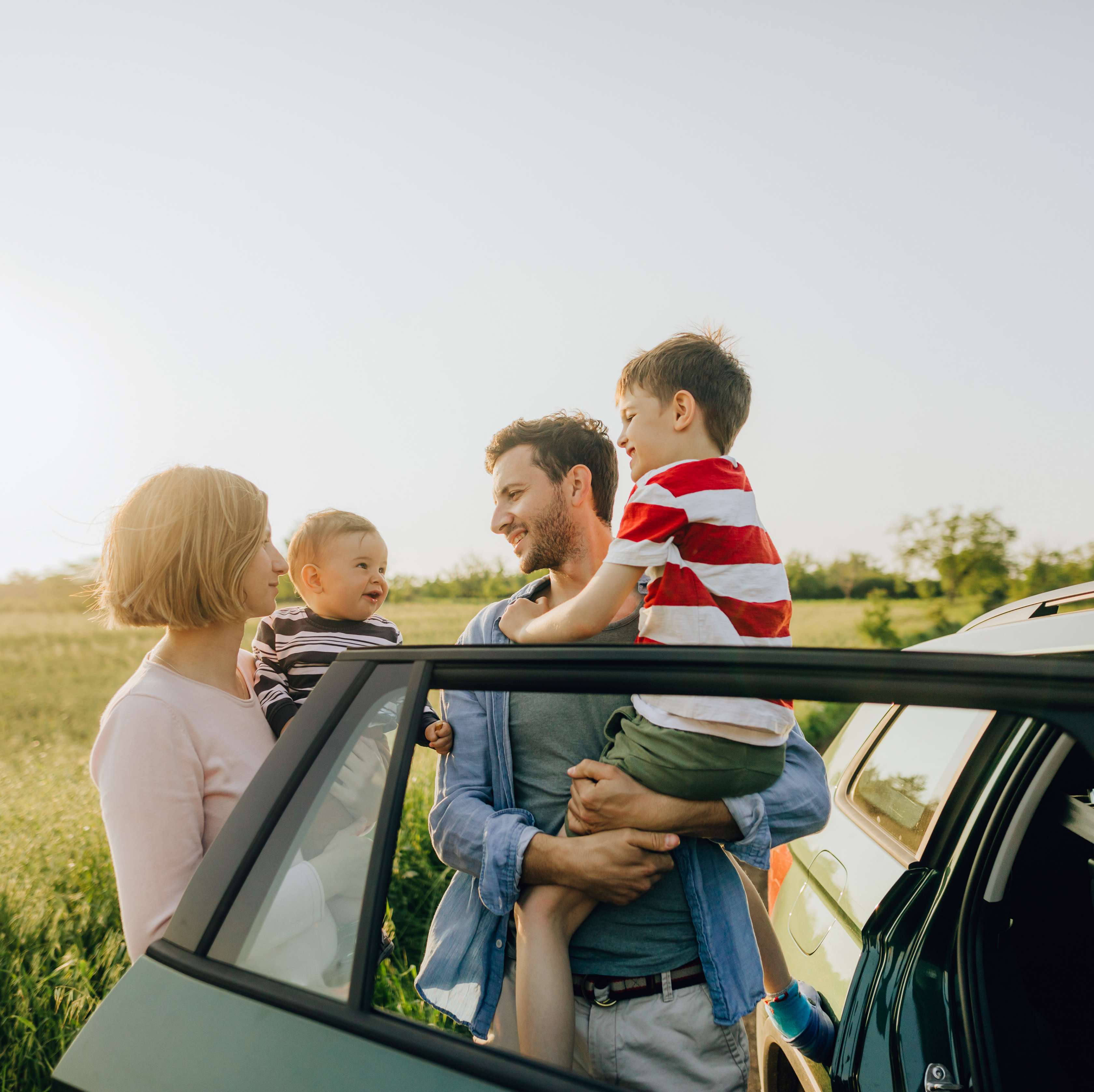 Mann und Frau halten zwei kleine Kinder im Arm, während sie vor ihrem Auto auf einem Feld stehen.
