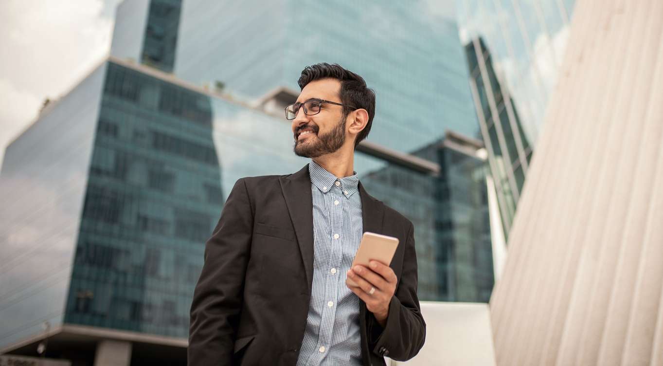 Meine Allianz | Dokumente Digital: Ein Geschäftsmann steht mit Handy in der Hand vor Bürogebäuden.