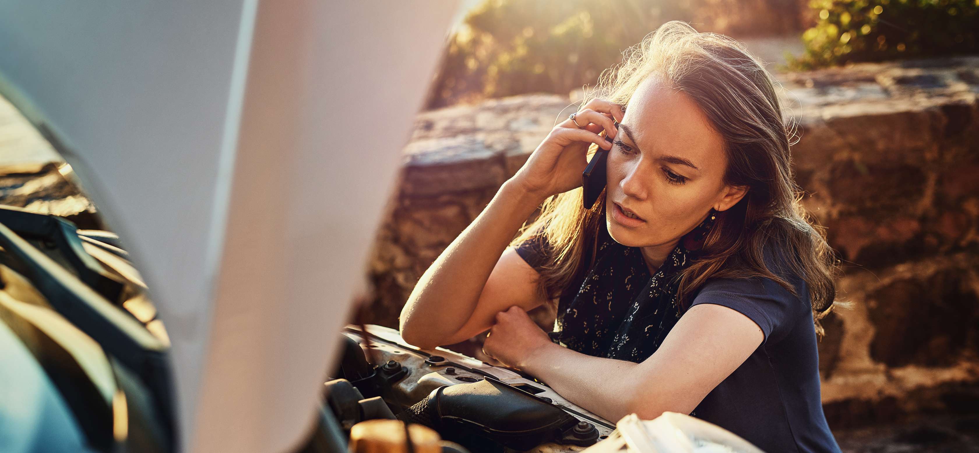 Allianz Kfz-Auslandsschaden melden: Eine Frau kniet vor der Motorhaube ihres Autos und telefoniert