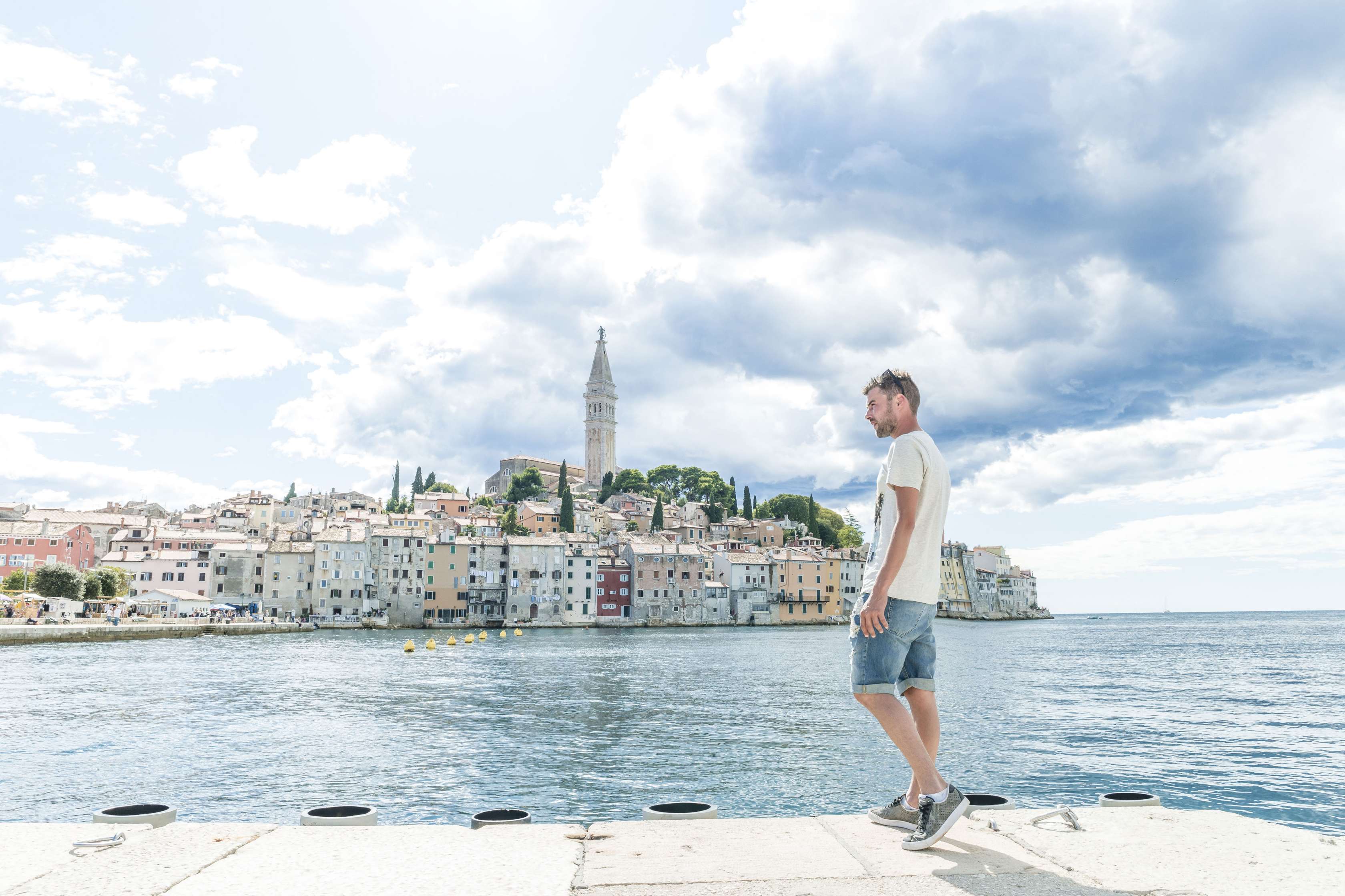 Mann steht am Meerufer, im Hintergrund ist eine Hafenstadt zu sehen