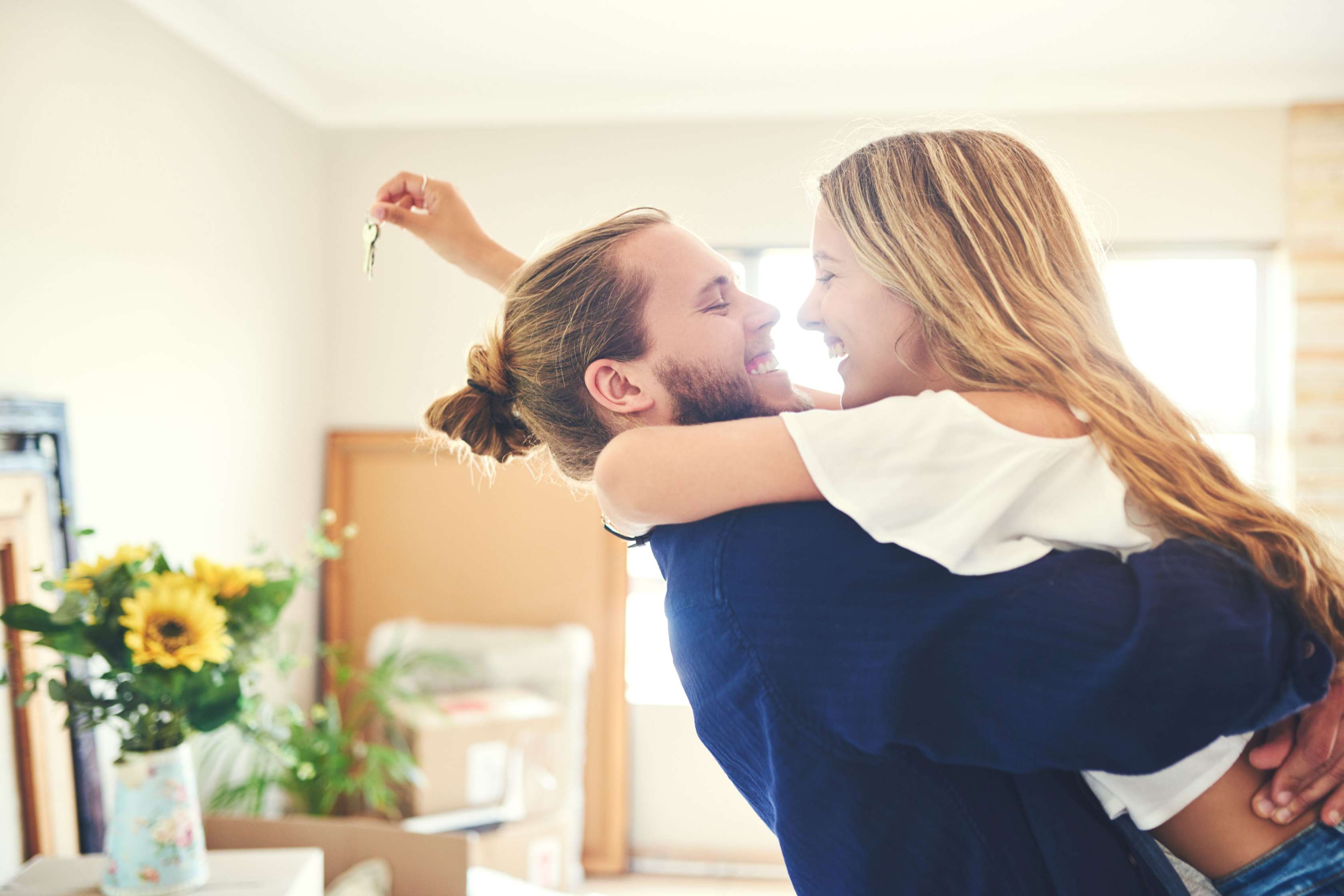 Ein junges Paar hält sich im Arm und lächelt sich fröhlich an. Im Hintergrund sieht man ihr neues Haus, sie hält den neuen Hausschlüssel in der Hand.