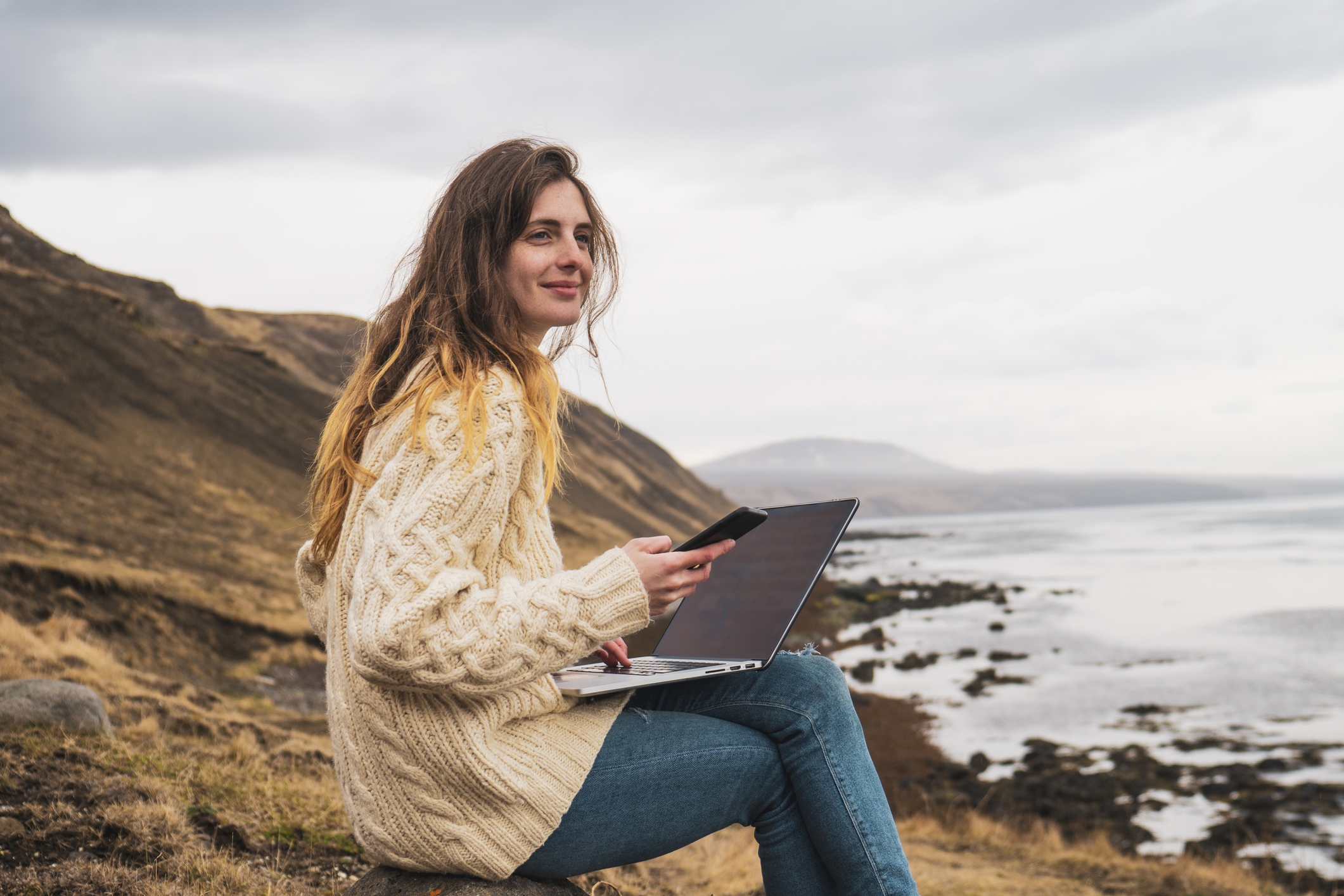 Frau sitzt mit Laptop an einer Küste und schaut zum Meer hinaus