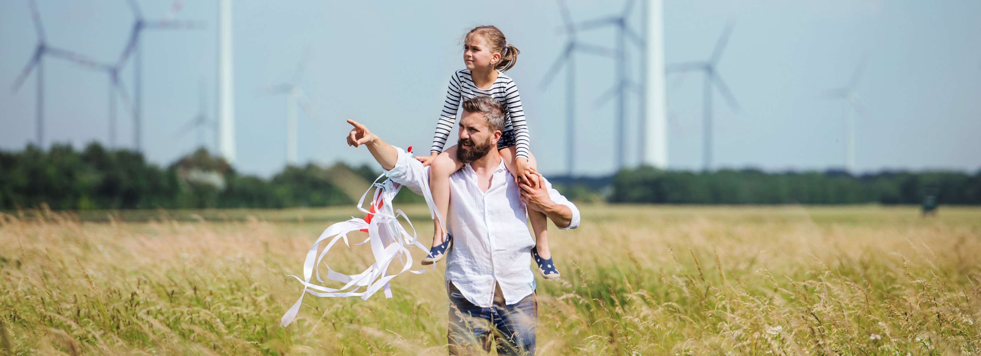 Nachhaltigkeit bei der Allianz: Ein Erwachsener trägt ein Kind auf den Schultern durch ein hohes Grasfeld. Der Erwachsene zeigt mit dem Finger in die Ferne, während das Kind einen Drachen hält. Im Hintergrund sind mehrere Windräder zu sehen, die sich vor einem blauen Himmel drehen.