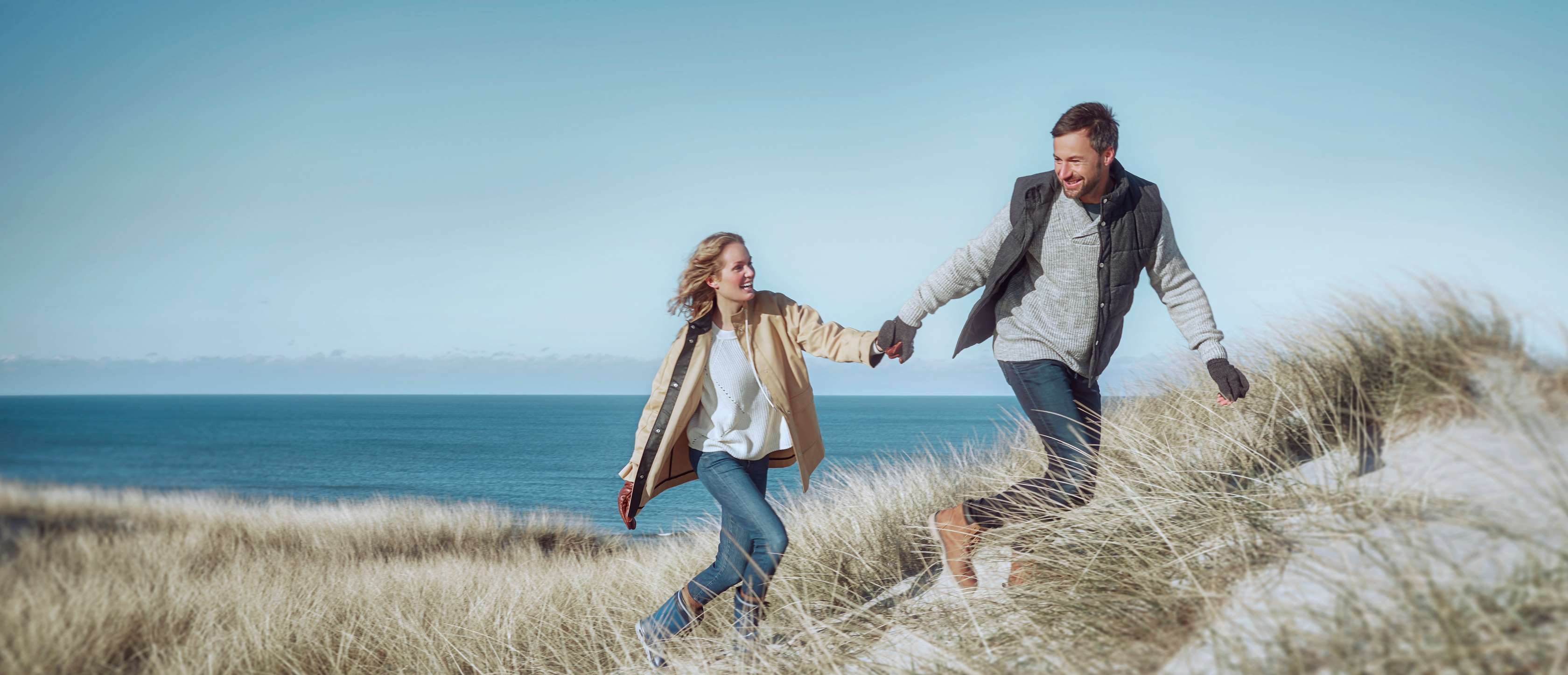 Allianz InvestFlex mit AktivDepots: Ein Paar hält sich an den Händen, während es lachend über grasbewachsene Dünen läuft