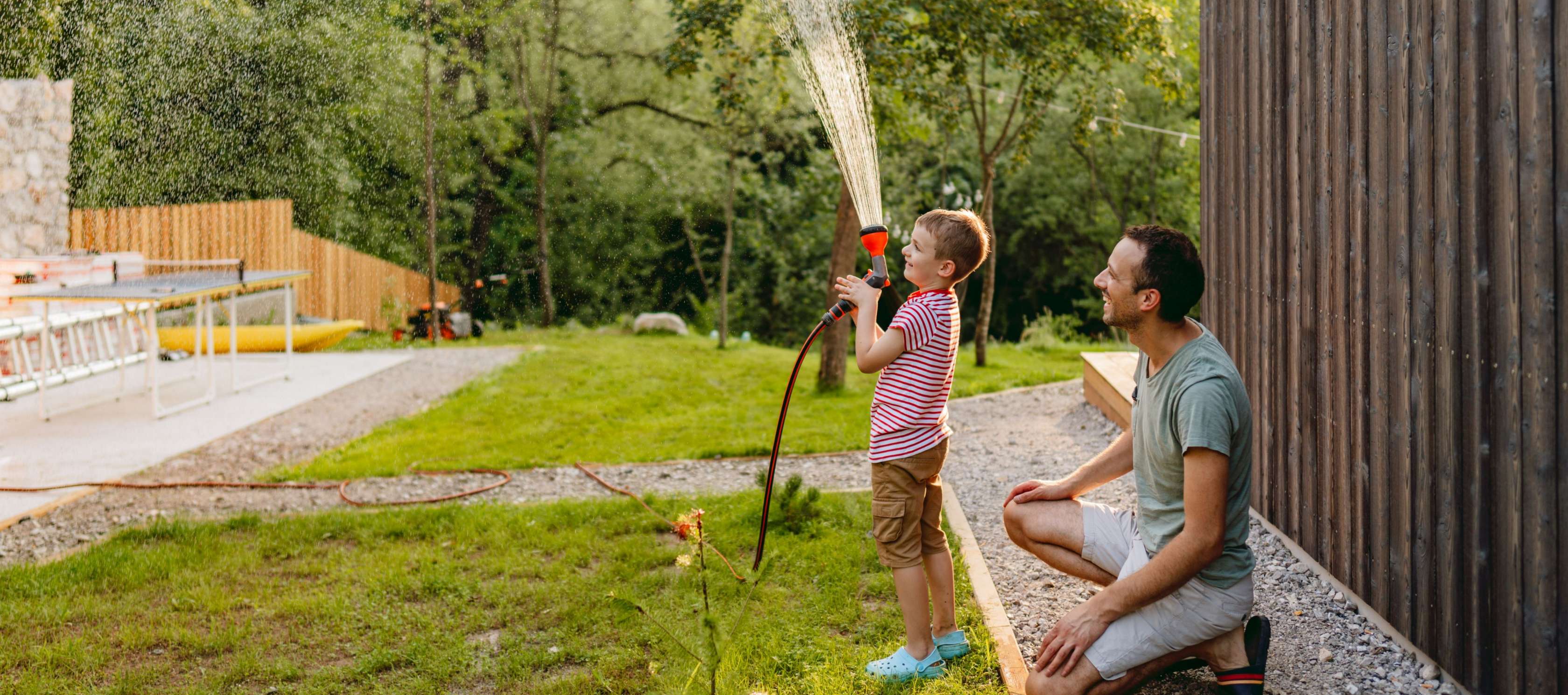 Allianz KinderPolice - Ein Vater steht im Garten mit seinem kleinen Sohn und sie gießen gemeinsam mit einem Schlauch den Garten.