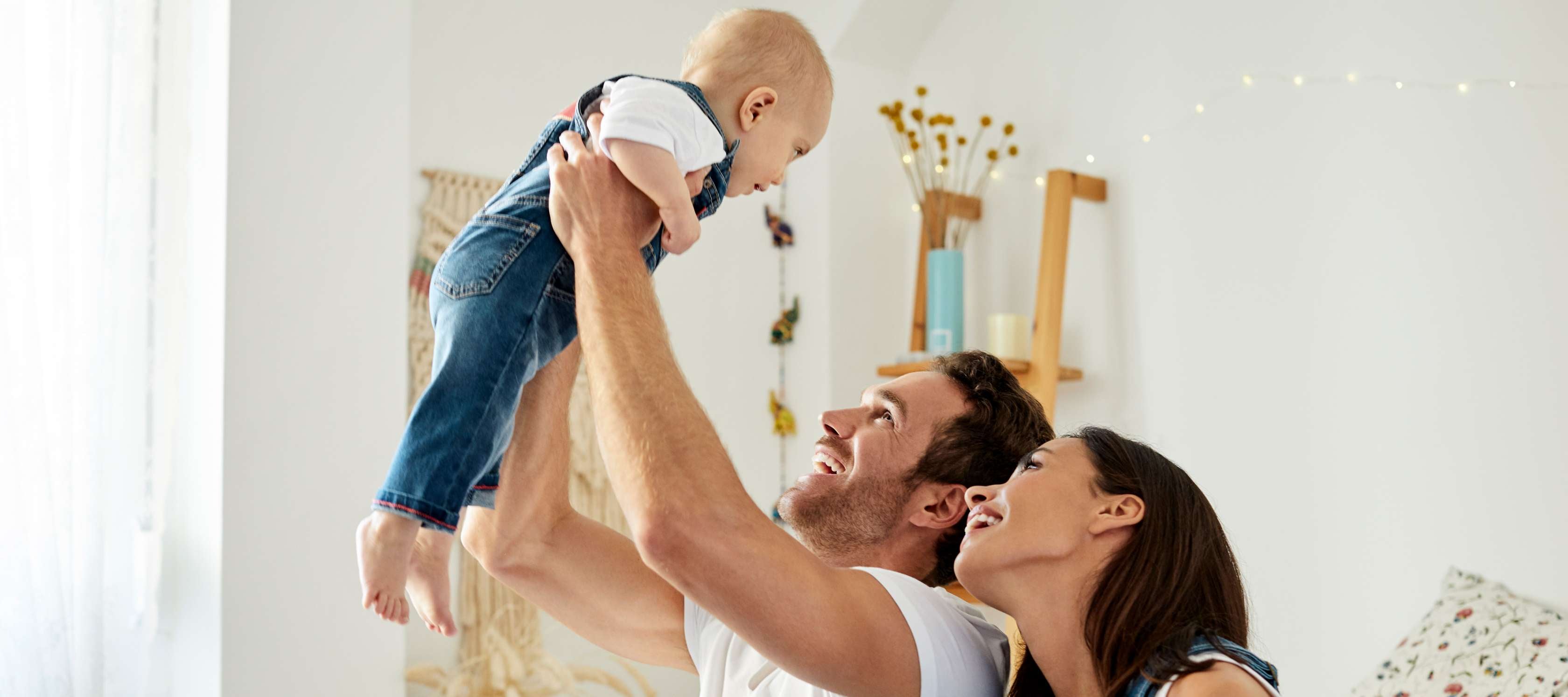 Allianz KinderPolice - Elternzeit: Ein Vater hält sein Kleinkind lächelnd in die Höhe, die Mutter steht daneben und schaut ebenfalls glücklich nach oben.