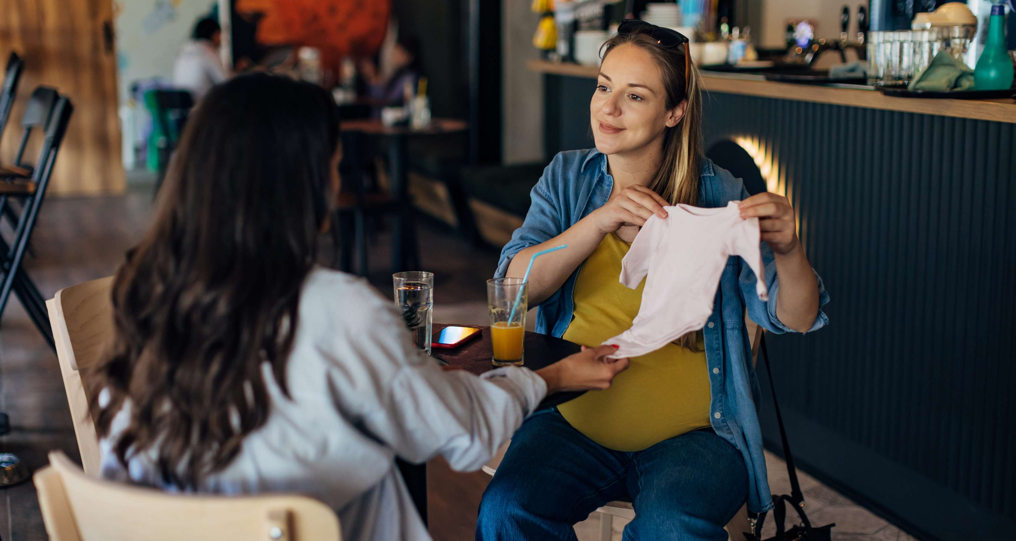 Allianz KinderPolice - Mutterschutz: Eine schwangere Frau hält lächelnd einen Baby-Body in der Hand, den sie einer Freundin zeigt.