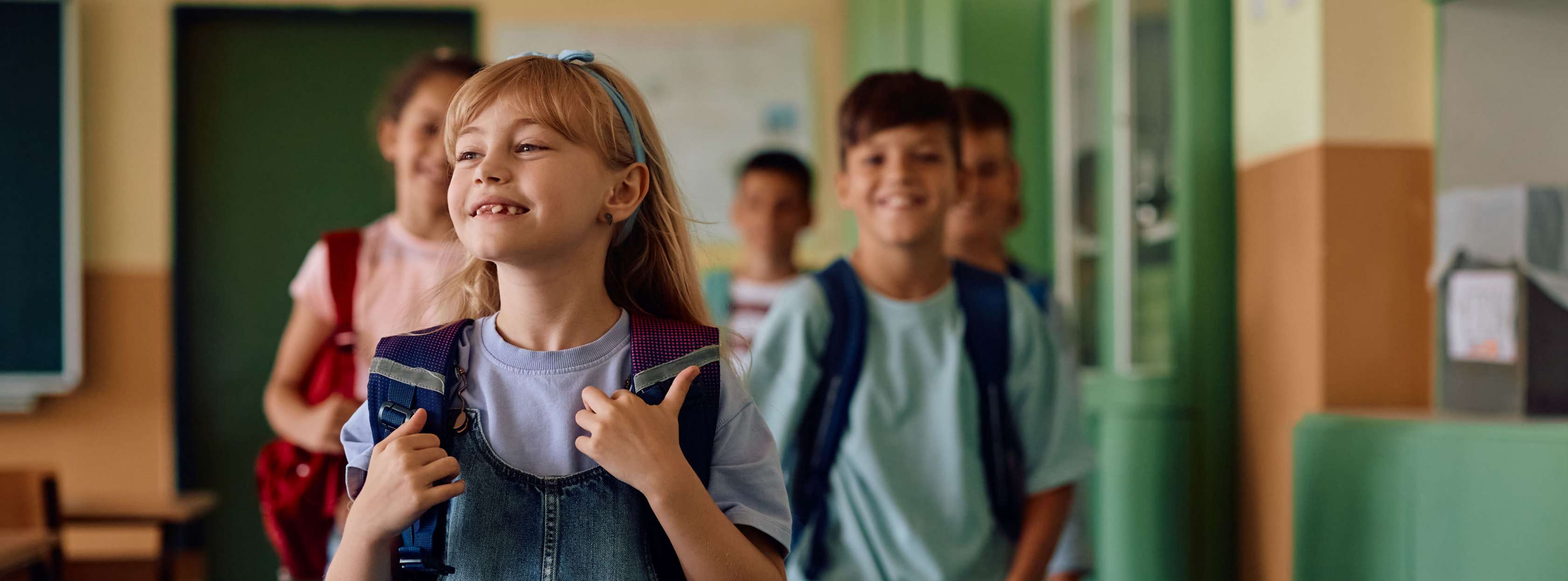 Allianz KinderPolice - Schulanmeldung Grundschule: Ein Mädchen läuft lächelnd durch den Schulfur, hinter ihr sieht man weitere Schulkinder, die glücklich sind.