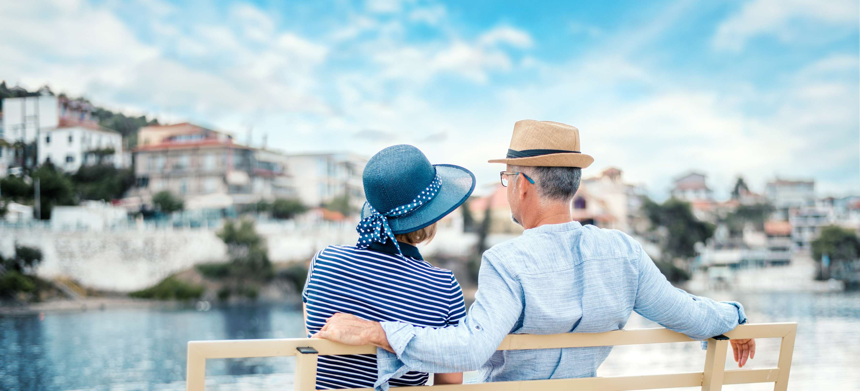 Allianz - Vorsorge - Ruhestand im Ausland: Älteres Paar in blauer Kleidung sitzt auf einer Bank und schaut zum Meer