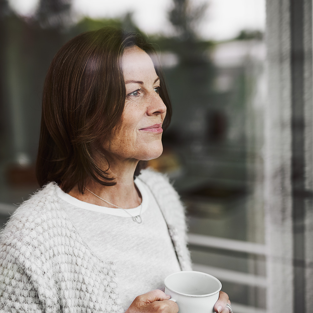 Eine Frau hält eine Teetasse in der Hand und blickt lächelnd und nachdenklich aus dem Fenster