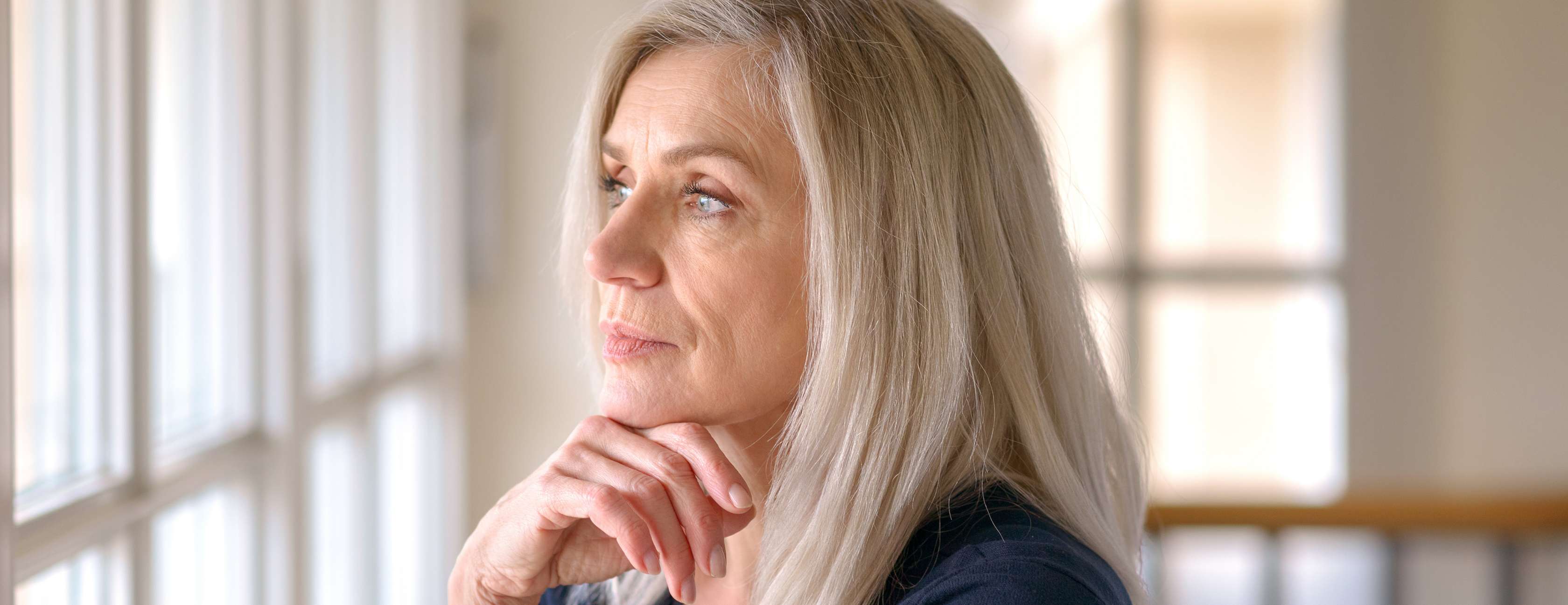 Allianz - Vorsorge - Rente aufstocken: Eine ältere Frau mit langen Haaren schaut nachdenklich aus dem Fenster.