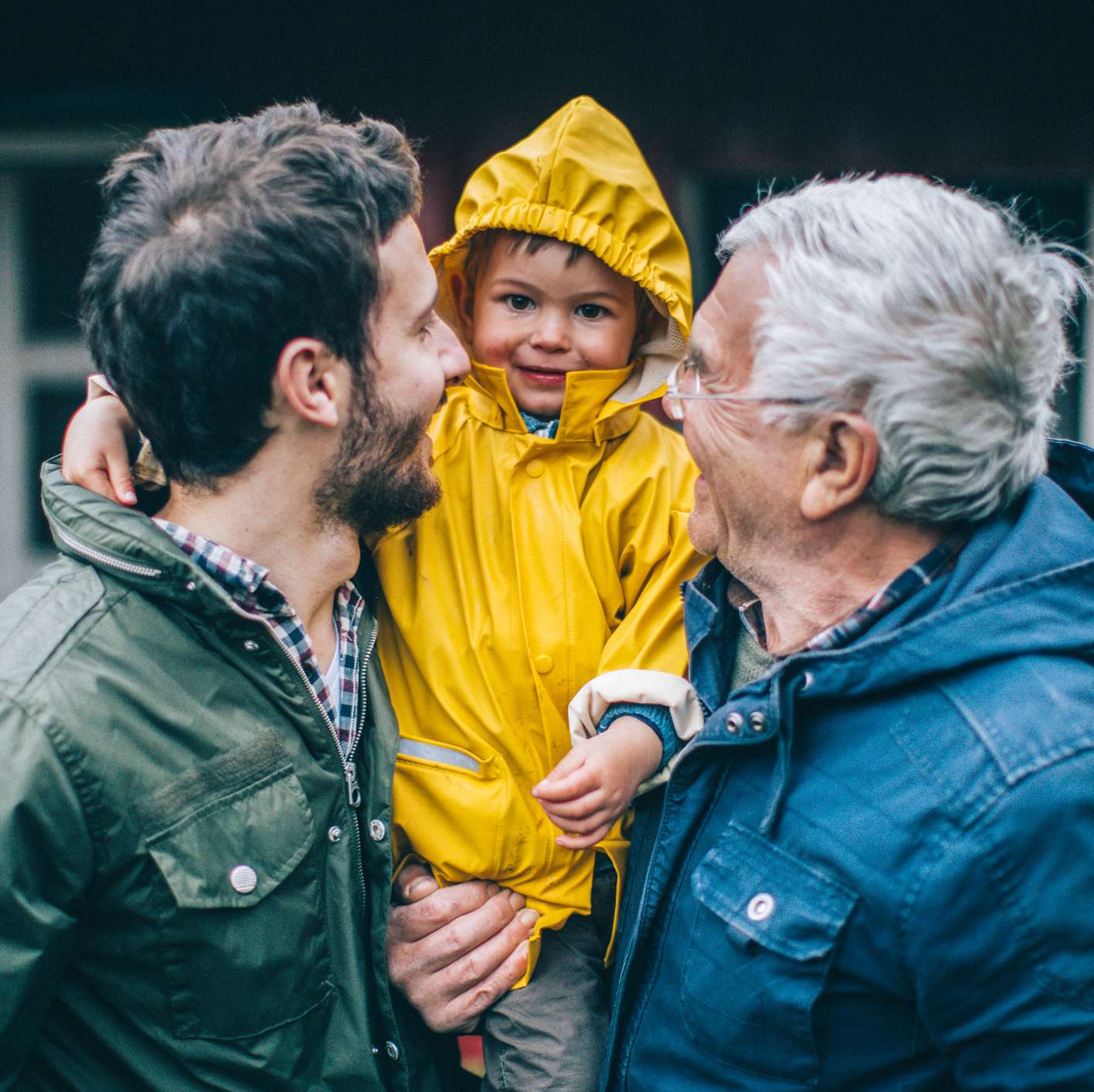 Allianz - Rentenwelt - Rente vererben: Man trägt Kleinkind im gelben Regenmantel auf dem Arm, älterer Mann auf der anderen Seite des Kindes lacht