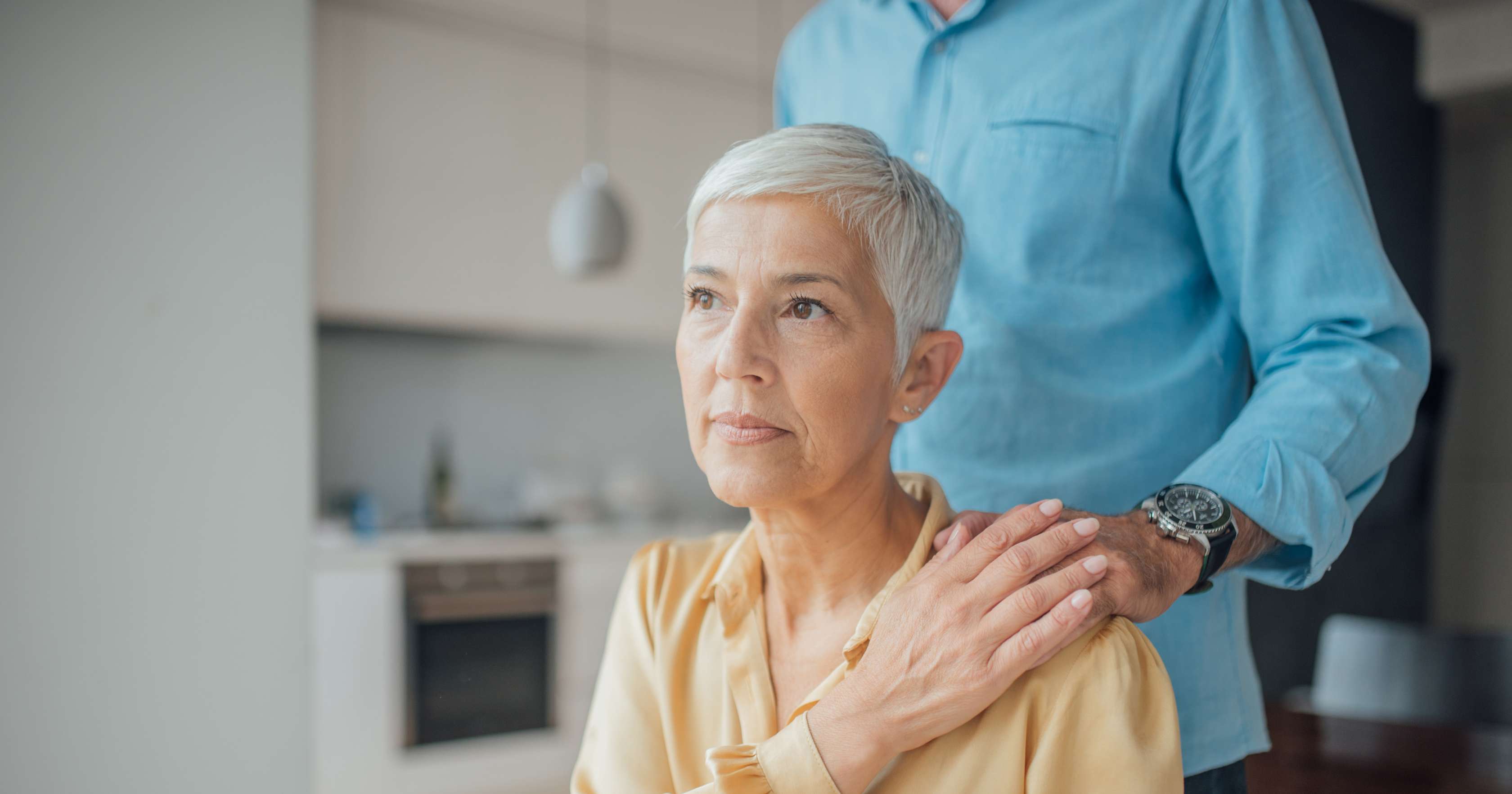 Allianz - Rente - Vererben: Mann steht hinter einer älteren Frau und legt ihr die Hand auf die Schulter