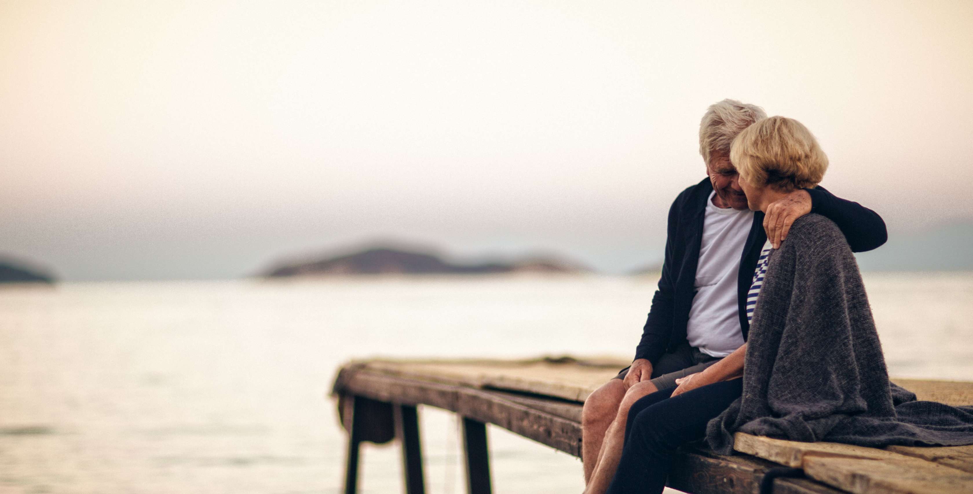 Allianz Berliner Testament - Ein altes Paar sitzt am Strand und legt den Kopf aneinander.