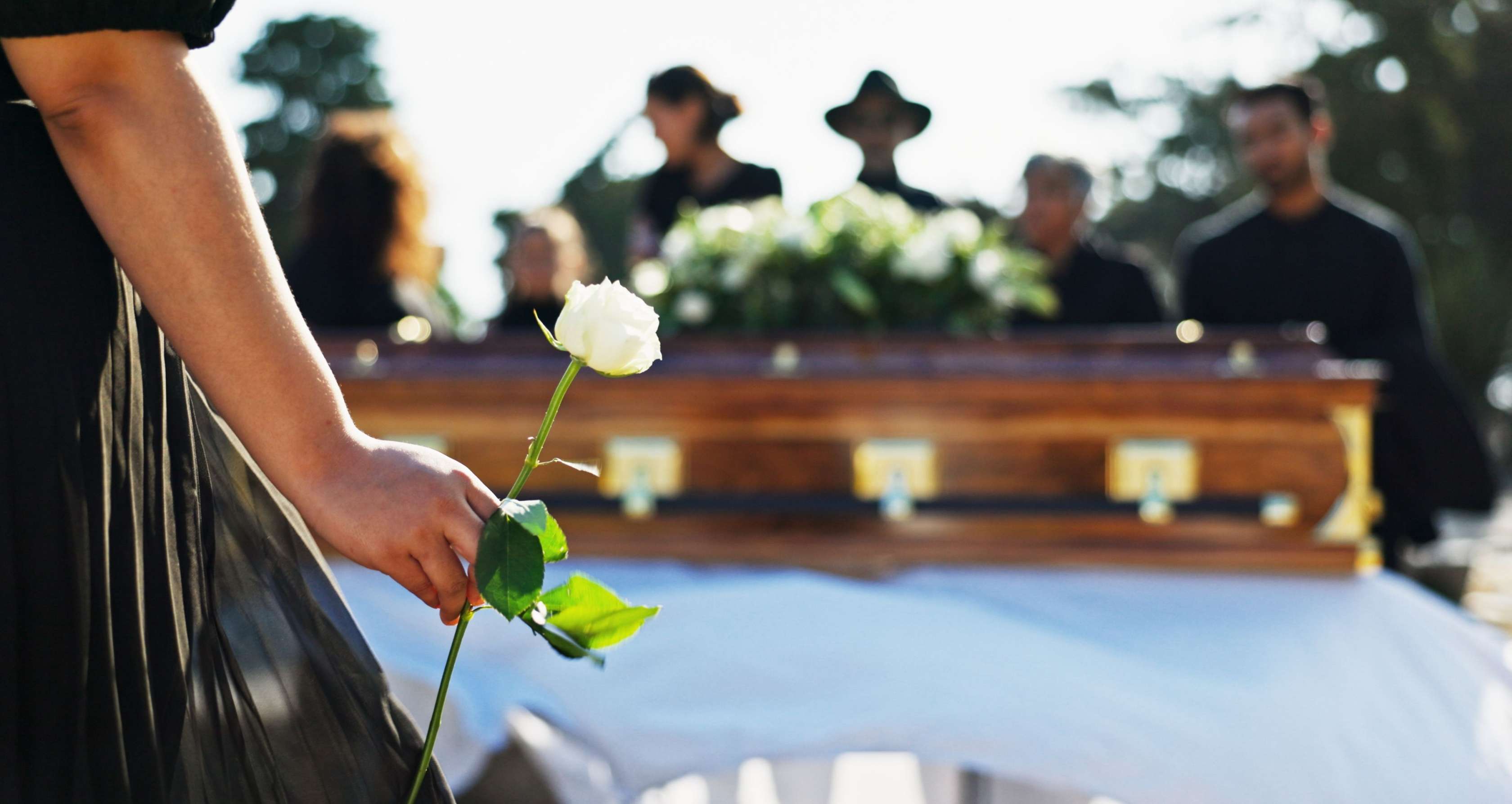 Allianz Sterbegeldversicherung - Beerdigungskosten absetzen: Im Vordergrund hält eine Frau eine weiße Rose in der Hand, im Hintergrund finde eine Beerdigung statt.