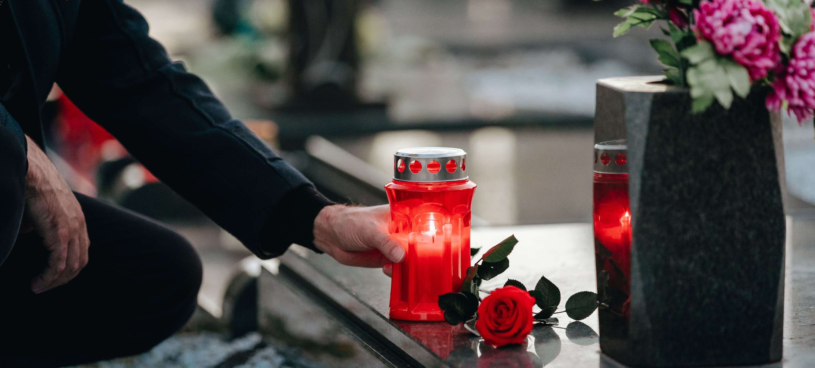Allianz Sterbegeldversicherung - Grabstein Kosten: Ein Mann stellt eine brennende Kerze auf einen schwarzen Grabstein., der mit Blumen geschmück ist.