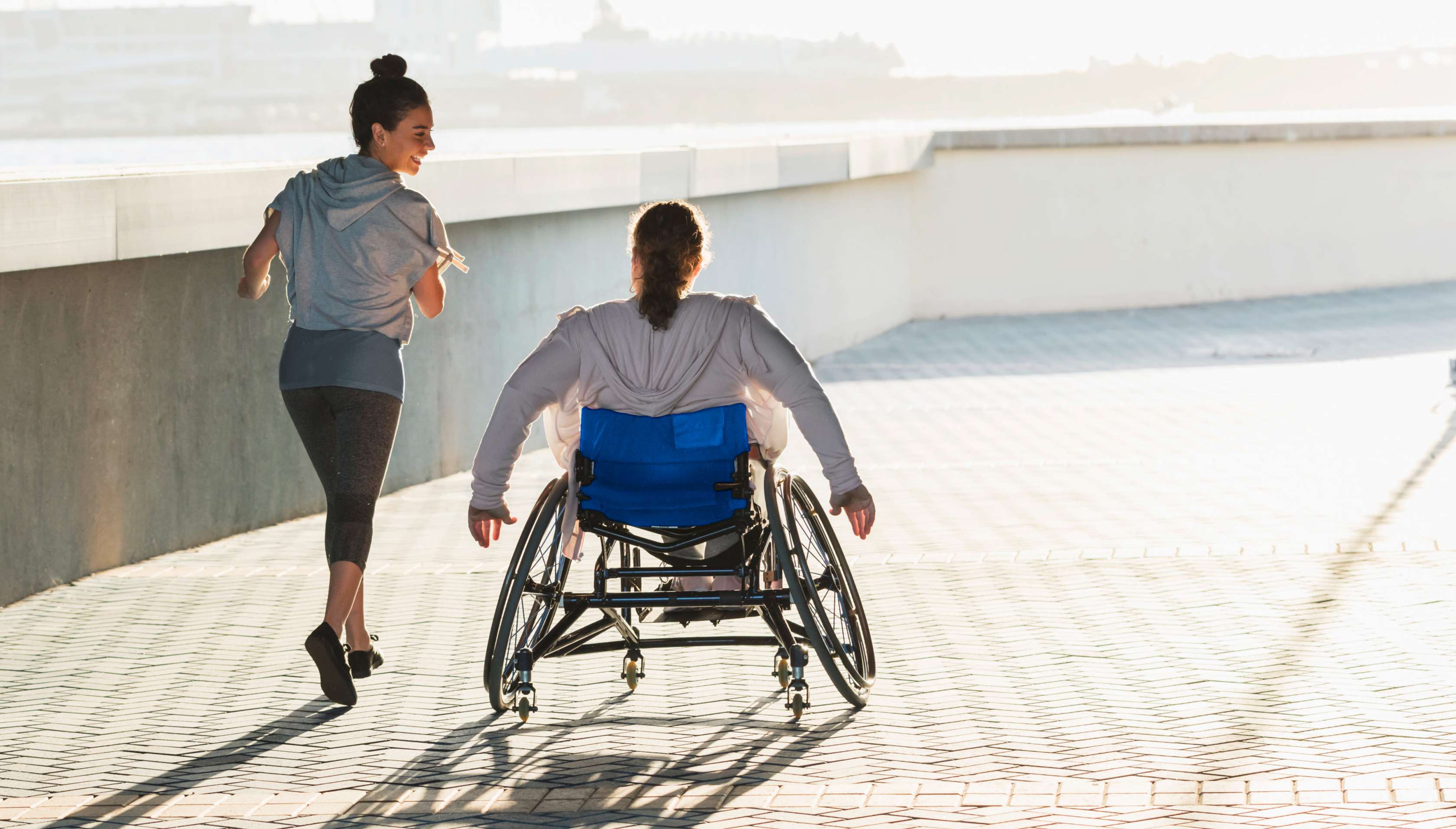 Allianz Unfallversicherung: Ein Rollstuhlfahrer fährt neben einer Joggerin auf einem Weg