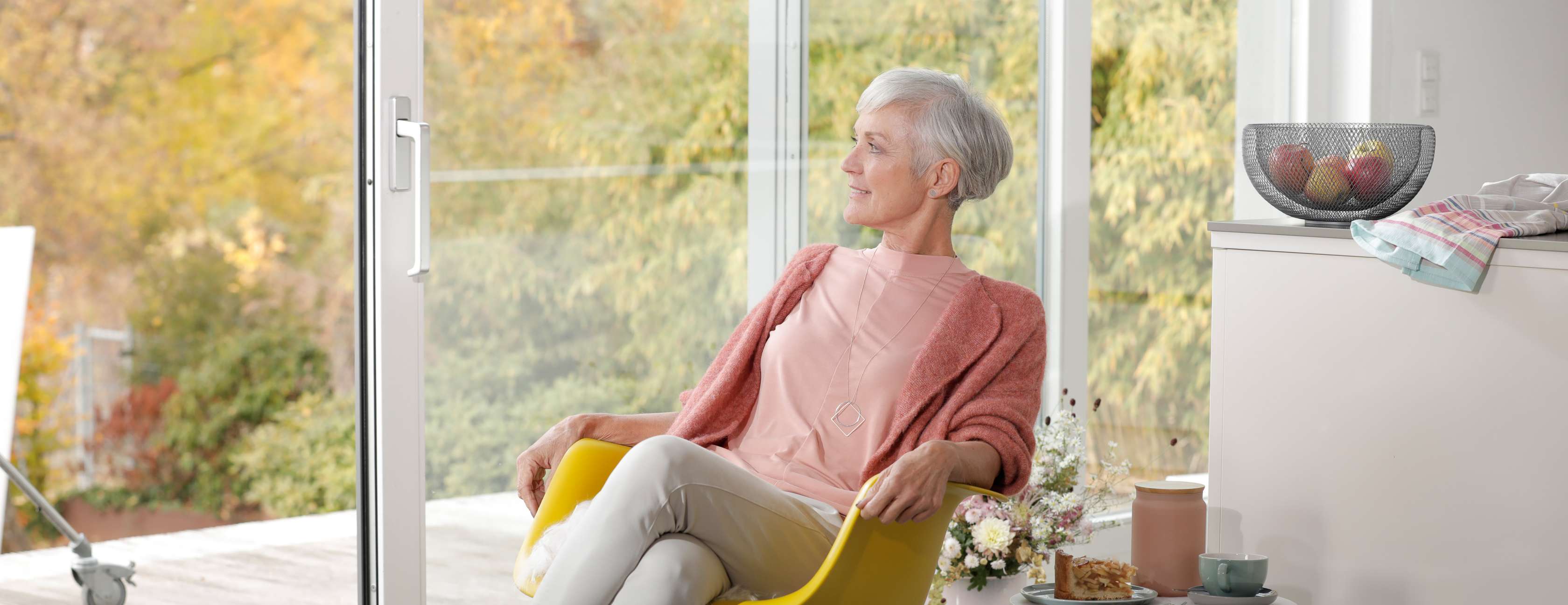 Ältere Frau sitzt entspannt in ihrem Wohnzimmer und schaut durch eine Schiebetür auf die Terrasse