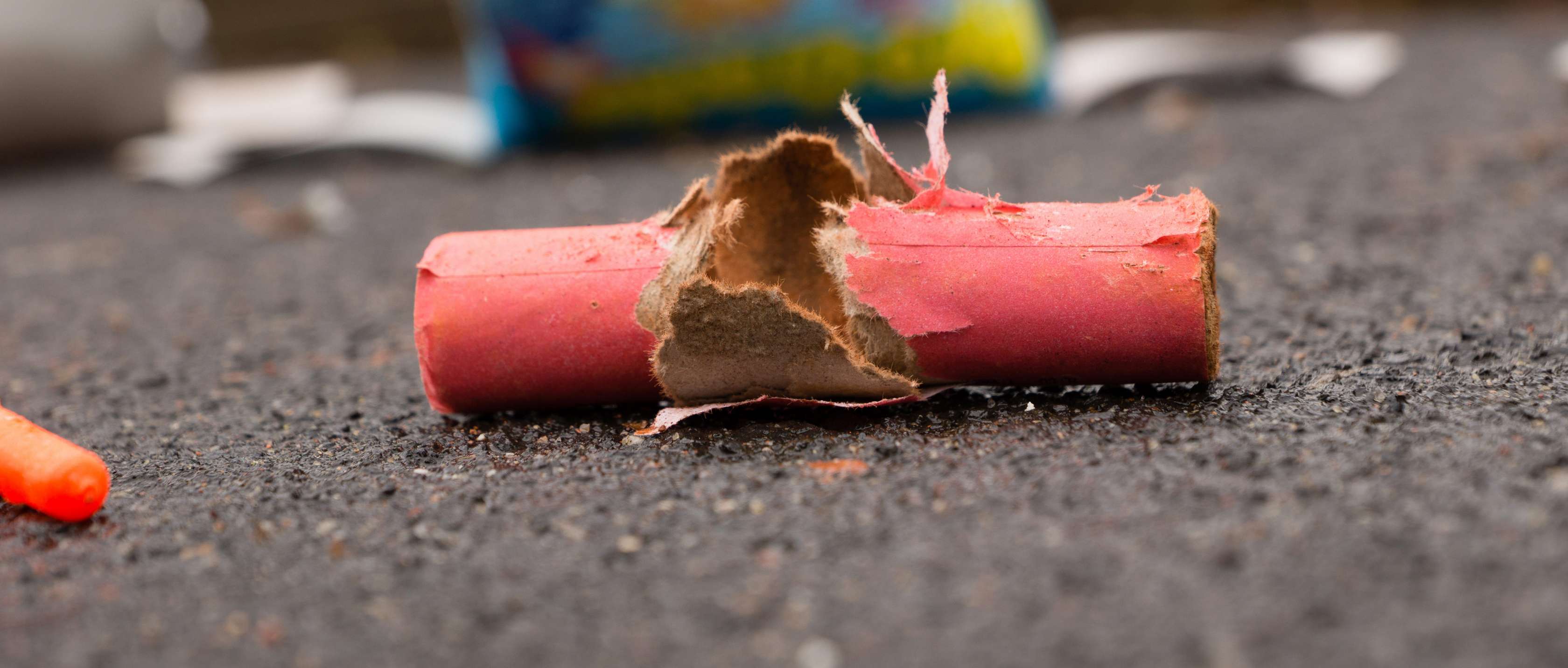 Allianz Unfallversicherung - Feuerwerksunfall: Roter Böller liegt zerrissen auf der Straße, im Hintergrund liegen Feuerwerkskörper.