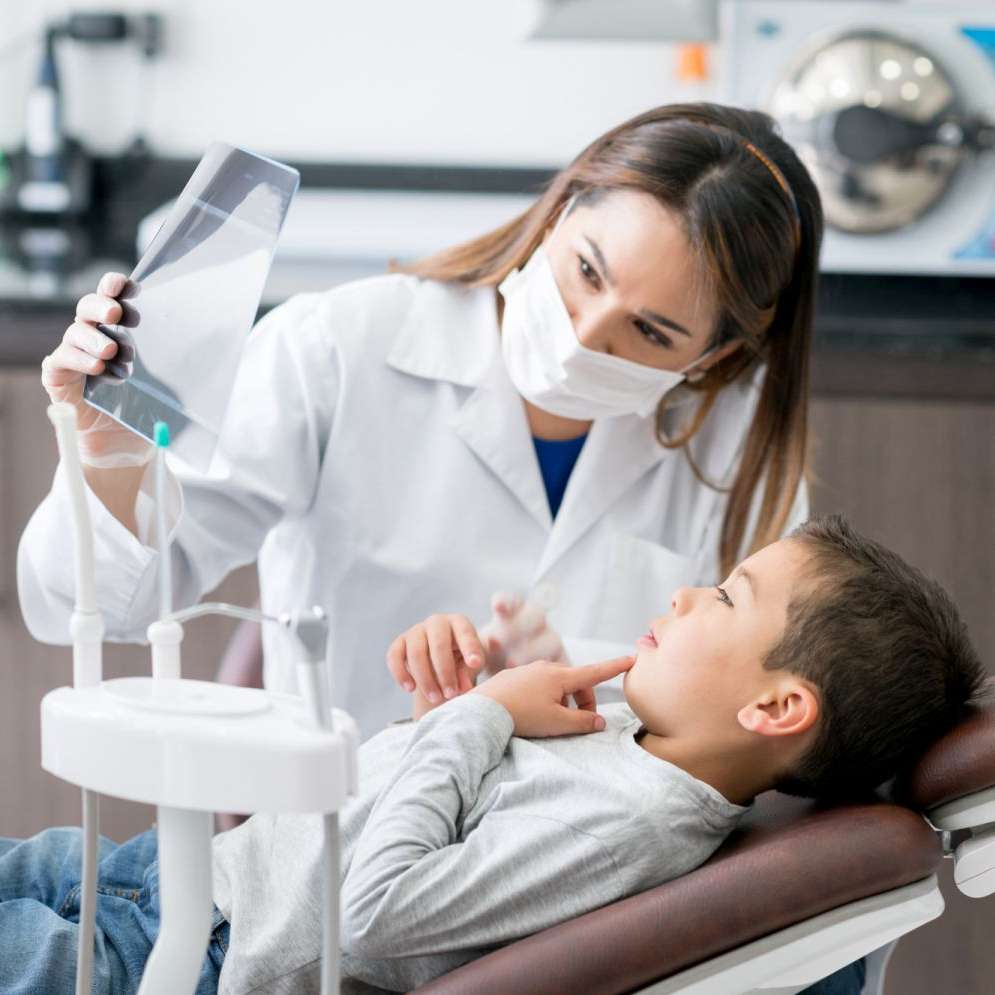 Zahnärztin mit Mundschutz schaut sich mit einem Jungen auf Behandlungsstuhl eine Röntgenaufnahme an